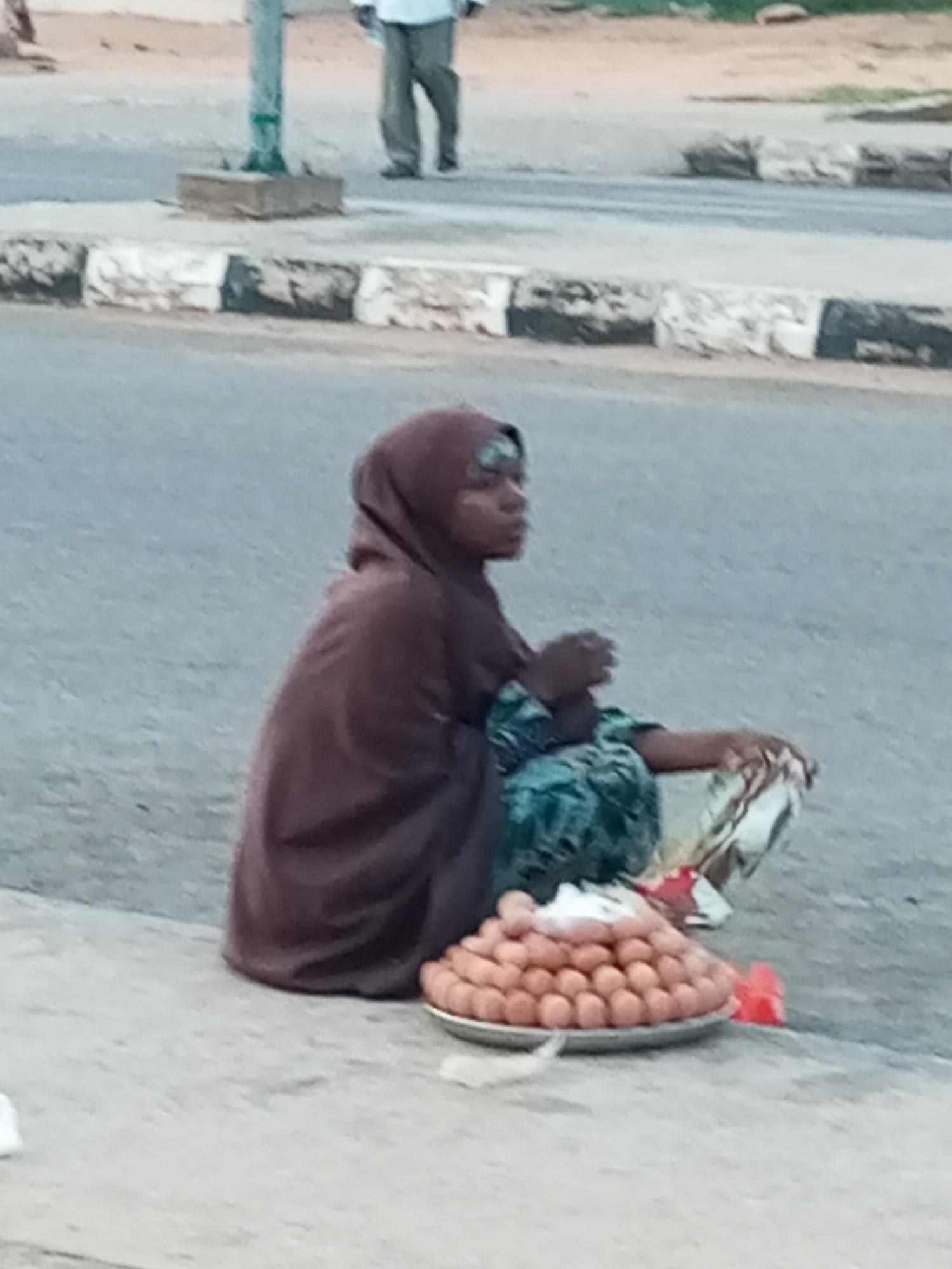 In Gombe, Nigeria, a woman wearing a brown hijab and a patterned dress sits on the side of a paved road. She is positioned next to a large plate piled high with what appear to be eggs. Her hands are resting on the plate. In the background, a person in a white shirt and gray pants walks away from the camera. The street has a curb with black and white markings.