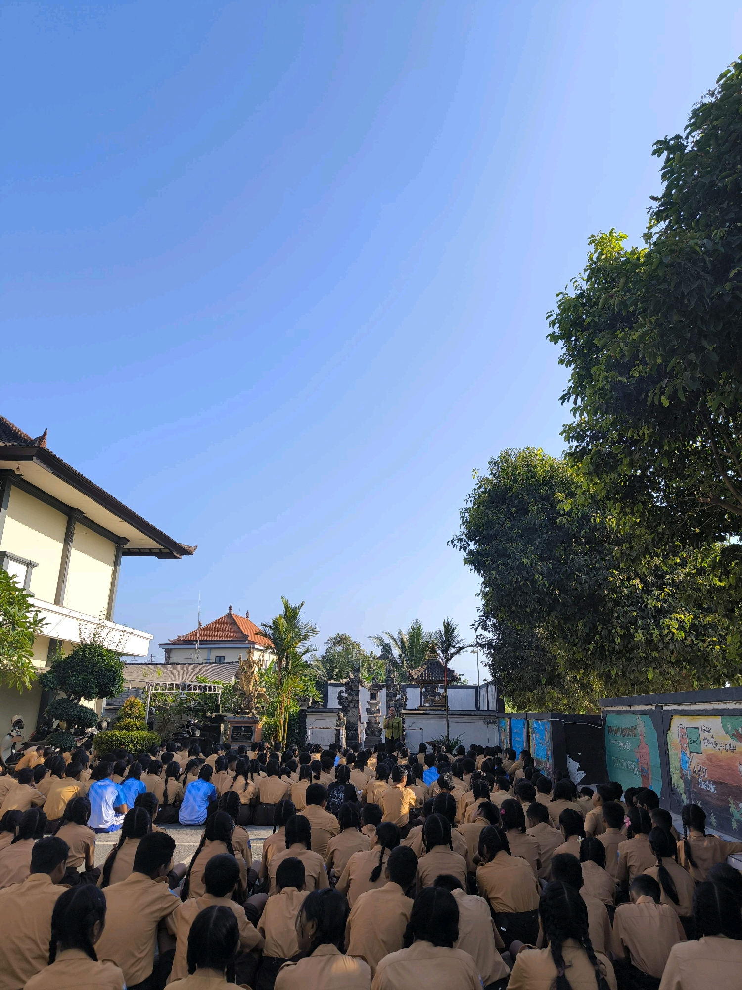 A group of students, dressed in light brown uniforms and some in blue shirts, are sitting on the ground facing a stage or platform in what appears to be a school courtyard. The students are arranged in rows, with many in the foreground and more receding into the background. A tree is visible on the right side of the image, and a building with a tiled roof is on the left. The sky is clear and blue. The overall scene suggests an outdoor assembly or event. Based on architectural style, such as the tiled roofs and decorative elements on the building, and the presence of tropical trees, this location is likely in a tropical region. While the image itself does not explicitly state the location, the provided context indicates it is the city of Tabanan, Indonesia.