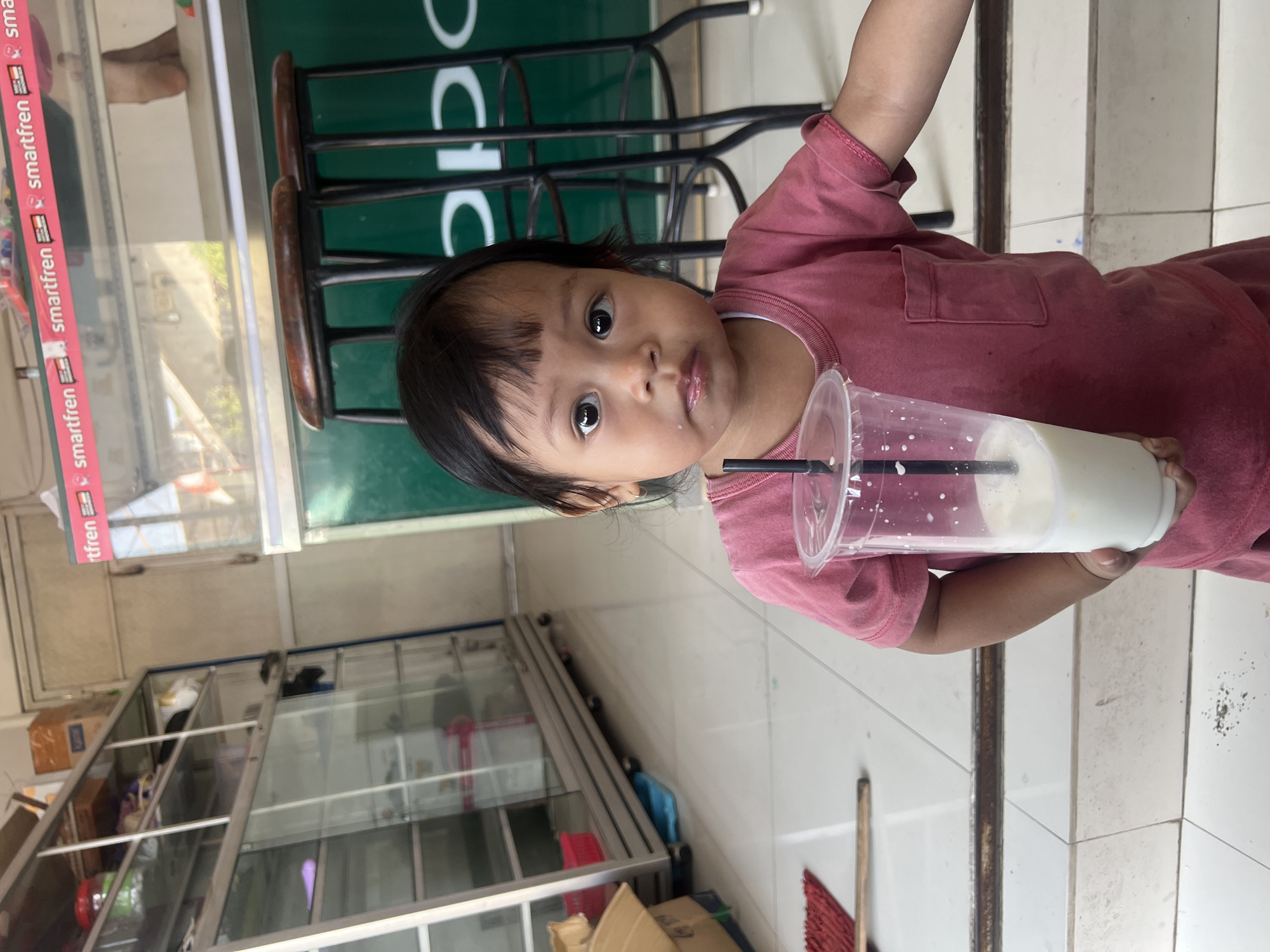 In a retail setting in Kecamatan Pati, Indonesia, a young child with dark hair and large, expressive eyes is holding a clear plastic cup filled with a white beverage and a black straw. The child is wearing a faded pink t-shirt. The background features a glass display cabinet with shelves, a green counter with stools, and advertisements for "smartfren" and "OPPO". The floor is tiled.
