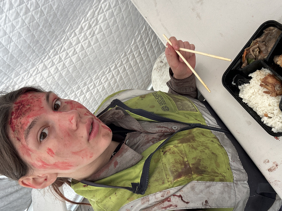 A young female subject is depicted within a temporary structure in Huangdao District, China. Her face, encompassing the forehead, cheeks, nose, and chin, is heavily covered with a vivid red substance consistent with blood, appearing as both smears and clots. Additional reddish-brown stains, indicative of dried blood and dirt, are present on her neck, a grey collared shirt, and a lime green and white high-visibility vest. The vest features reflective strips, a black zipper, and the logo "UEG-ESCO" with a building icon. The subject holds a pair of wooden chopsticks in her right hand, positioned above an open black, compartmentalized food container. The container holds white rice, a portion of fried poultry or similar item, and a beef and vegetable dish. The subject is seated at a white, flat surface, and the immediate background consists of a silver, quilted, insulated material, indicative of a tent or temporary shelter wall. She maintains direct eye contact with the viewer, exhibiting a neutral facial expression.