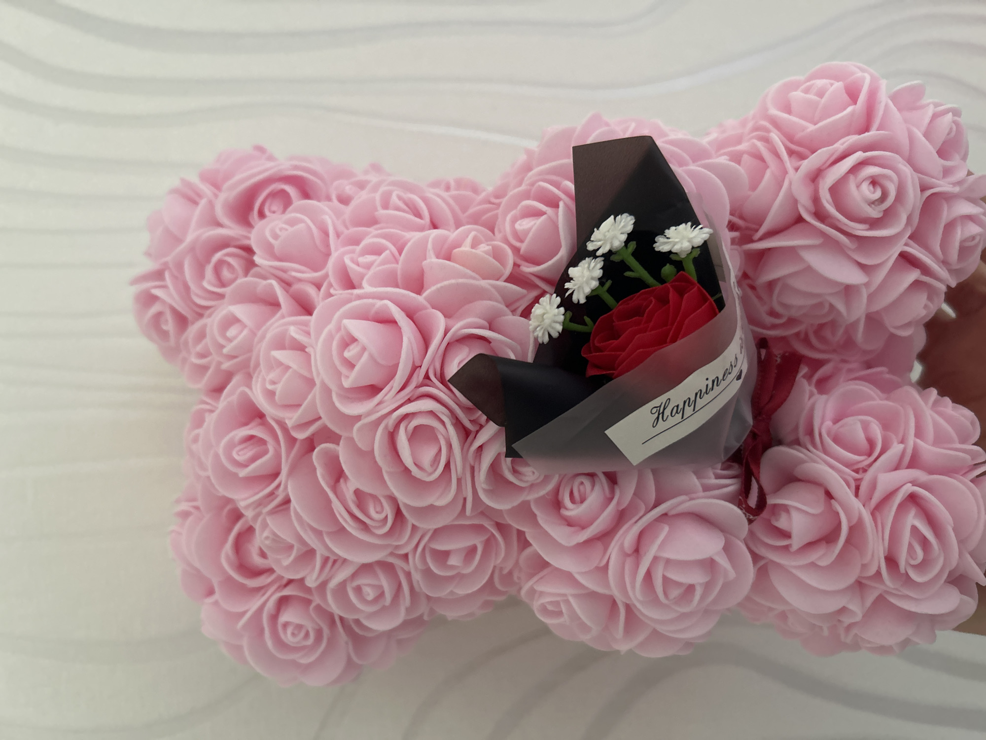 A close-up shot shows a pink rose teddy bear with a small bouquet of flowers on top. The bouquet consists of a single red rose, several small white flowers with green stems, wrapped in black paper with a clear plastic overlay, and tied with a red ribbon. A tag that reads "Happiness" is attached to the bouquet. The teddy bear is made entirely of pink roses. The background is a white surface with a light, wavy pattern. No location is specified, and there is no mention of Strasbourg, France.