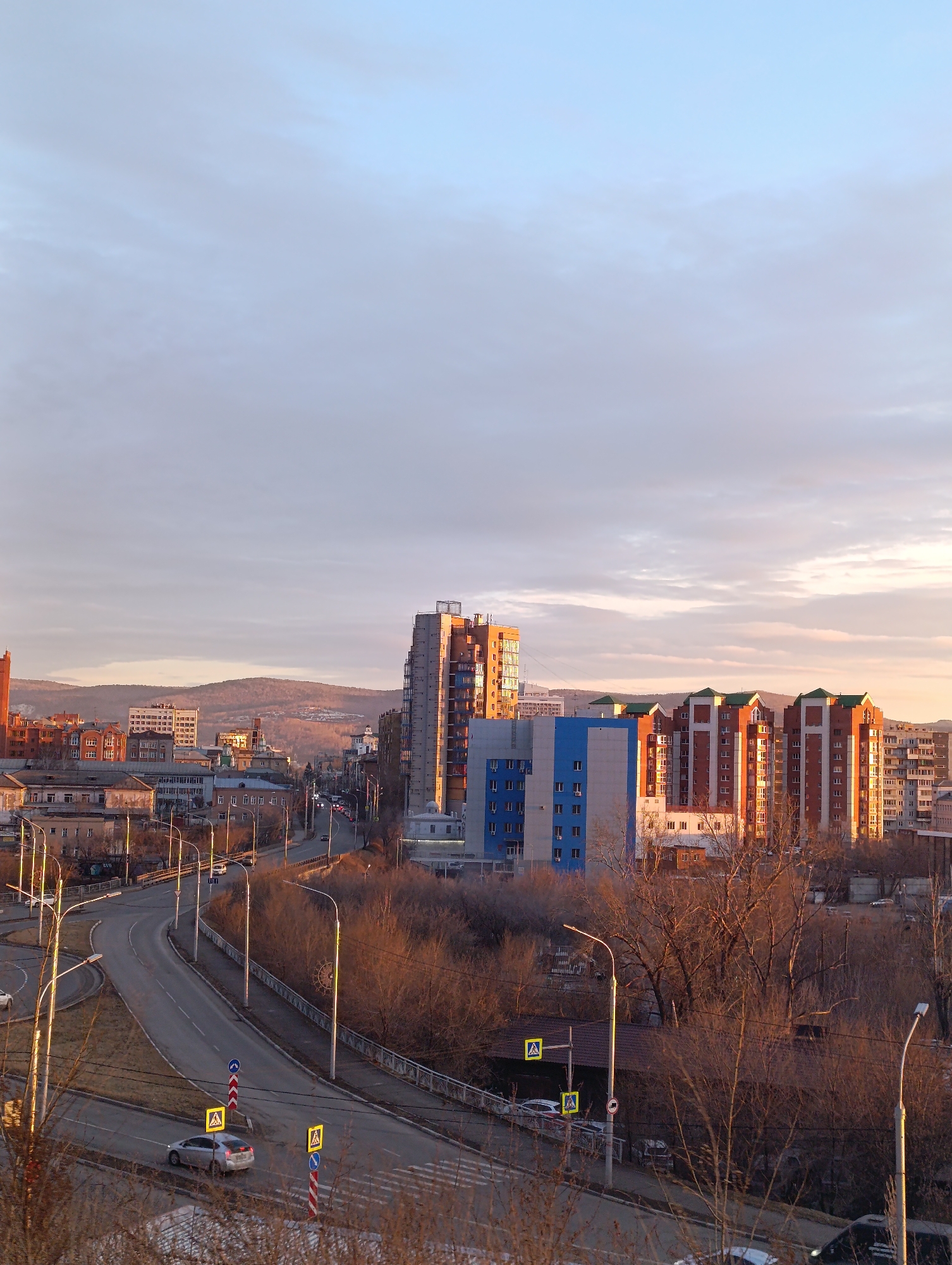 The image depicts a cityscape in Krasnoyarsk, Russia, taken from an elevated perspective. The scene is bathed in the warm light of either sunrise or sunset.
**Environment:**
The sky is a blend of pale blue and soft gray clouds. The terrain is characterized by rolling hills in the distance.
**Cityscape:**
Dominating the urban landscape are residential buildings of varying heights and architectural styles. A prominent high-rise building with a facade reflecting the sunlight stands out. Another group of apartment buildings is clad in red brick with green roofs. A blue and grey building is also visible.
**Infrastructure:**
A multi-lane road curves through the foreground. Streetlights line the road. Pedestrian crosswalks are marked with yellow "caution pedestrian" signs.
**Objects:**
Cars are traveling on the road. Bare trees without leaves are in the midground, suggesting it may be autumn or early spring.