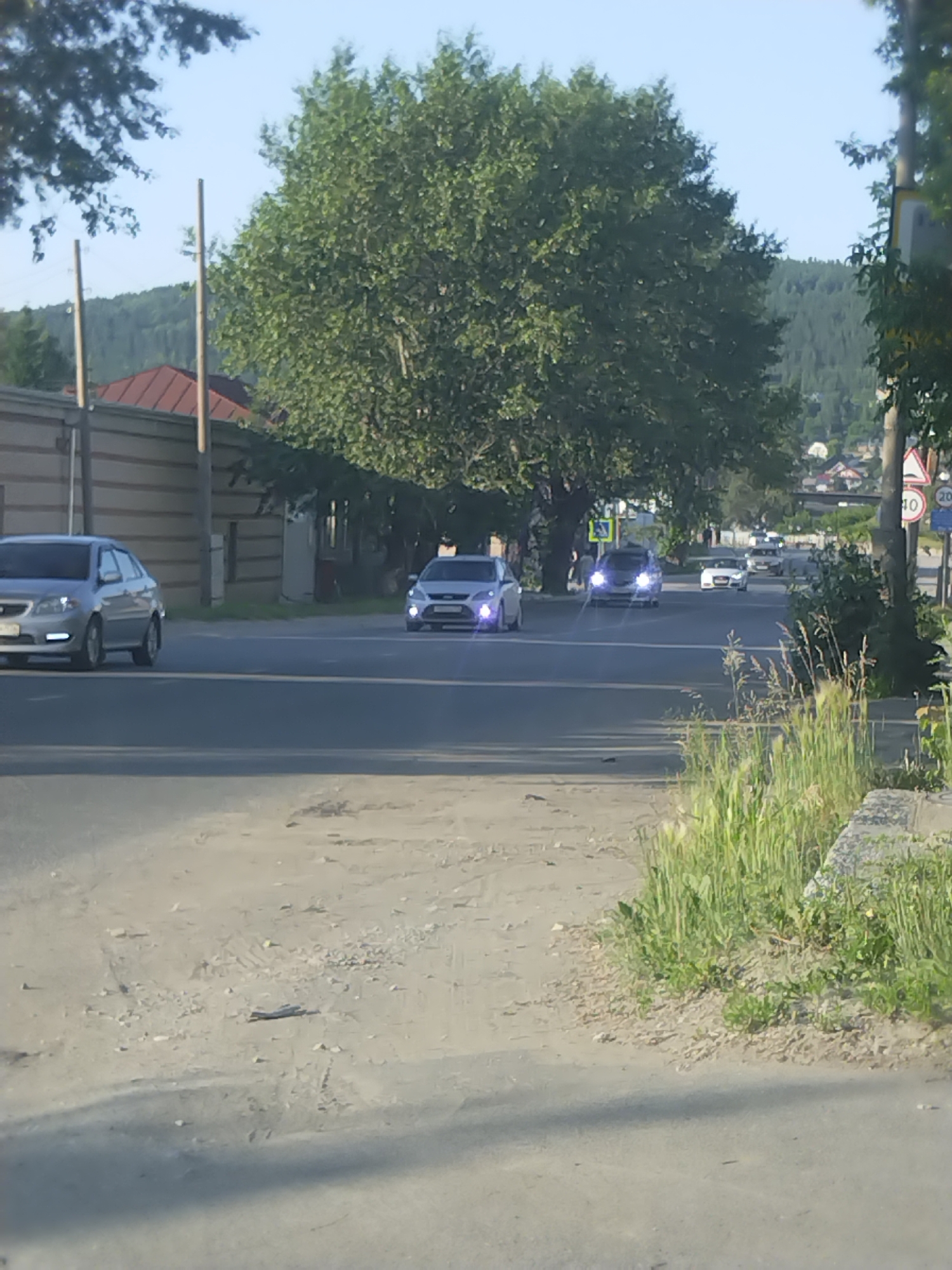 The image captures a street scene on a bright, sunny day, likely in a semi-urban or rural area, given the presence of trees, houses, and a road. The time of day appears to be late afternoon or early evening, judging by the long shadows cast on the road and the warm sunlight.
Several cars are visible on the road. On the left, a silver sedan is in motion. Further down the road, a white car with its headlights on is also moving. In the distance, more vehicles are present, indicating ongoing traffic. The road itself is paved, with some areas of dirt and gravel on the side, suggesting it may be a less-trafficked route or experiencing some construction or wear.
The background features lush green trees, including a very large, mature tree that dominates the center of the frame. Beyond the trees, hills covered in dense forest rise, and glimpses of houses with red roofs can be seen nestled amongst them, hinting at a residential area or village. The sky is clear and blue, contributing to the overall bright and pleasant atmosphere.
There are a few signs visible. On a pole on the right side of the road, there are traffic signs, including one that appears to indicate a speed limit, possibly "40" or "30". Another sign with a circle and a diagonal line is also present. A smaller blue sign with white text is also visible on the same pole.
The scene appears peaceful and ordinary, depicting everyday life with cars traveling on a road surrounded by nature and residential elements. There are no prominent human figures, but the presence of cars suggests human activity. The overall impression is one of a serene, sunlit day in a scenic location.