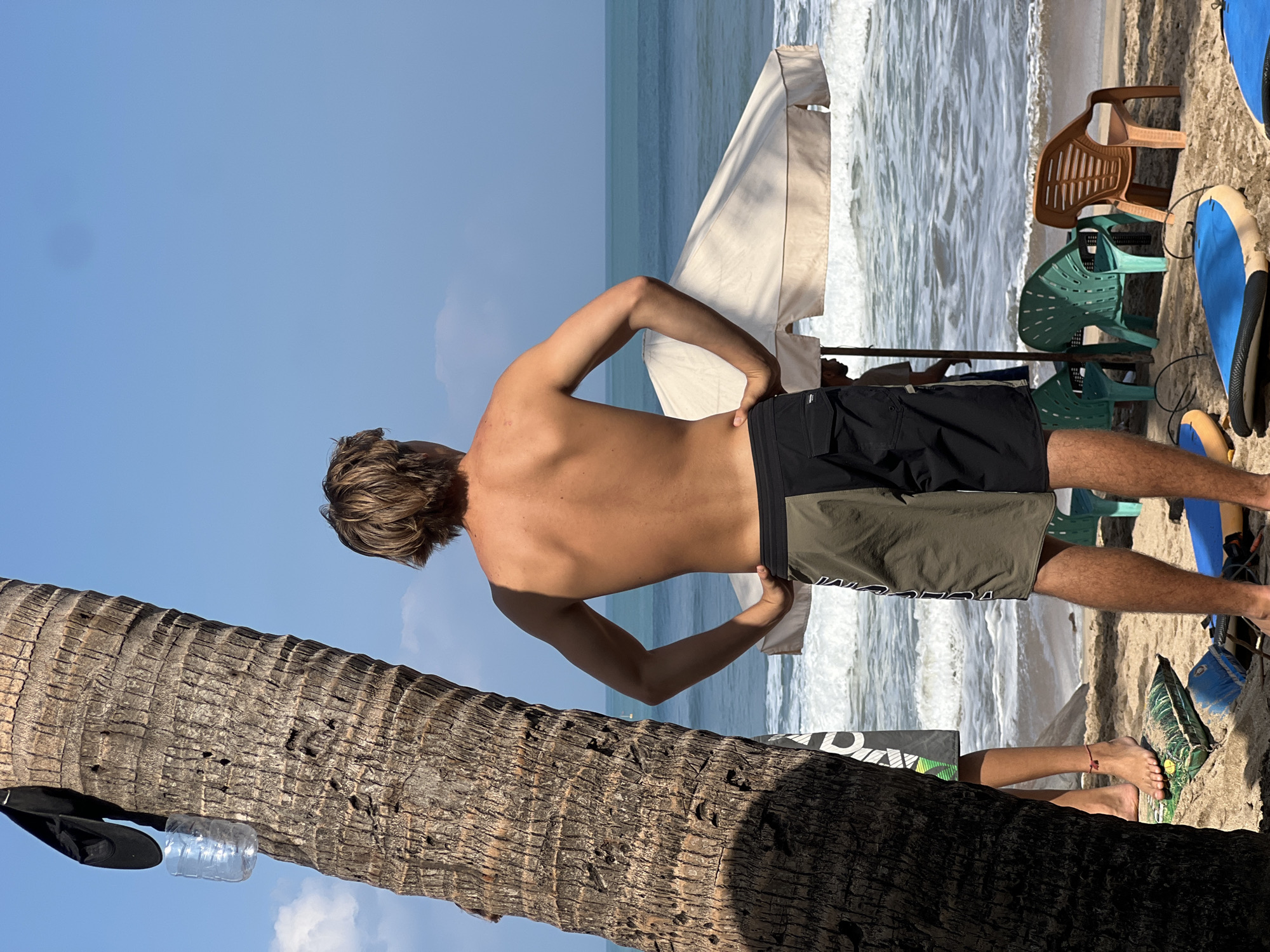The image depicts a beach scene in Kuta, Indonesia, with a young man standing in the foreground facing away from the camera. He has blondish-brown hair and is shirtless, wearing patterned board shorts. His hands are on his hips, and he appears to be looking out at the ocean. To his left, a large palm tree trunk dominates the frame, with a black cap and a plastic water bottle hanging from it. Behind the man, the ocean is visible with moderate waves breaking on the shore. There are beach chairs and umbrellas set up on the sand, and what appear to be surfboards in the foreground. Another person is partially visible behind the palm tree, wearing shorts. The sky is clear and blue.