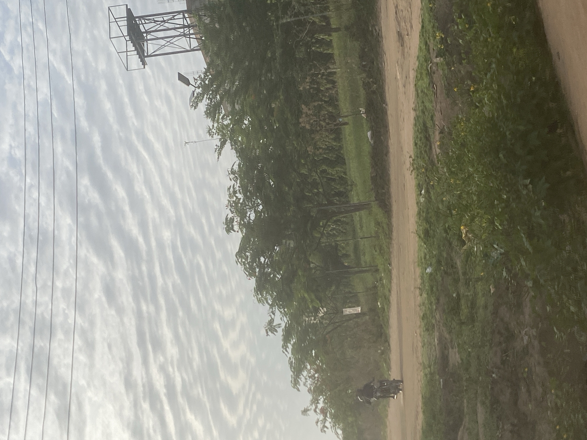 The image shows a dirt road in Iddo Tudunwada, Nigeria. The sky is covered with streaky clouds. Power lines are visible on the left side of the image. A metal utility tower is in the upper left corner. Trees line the left side of the road. A person rides a motorcycle on the road in the distance. Vegetation grows on the right side of the road.