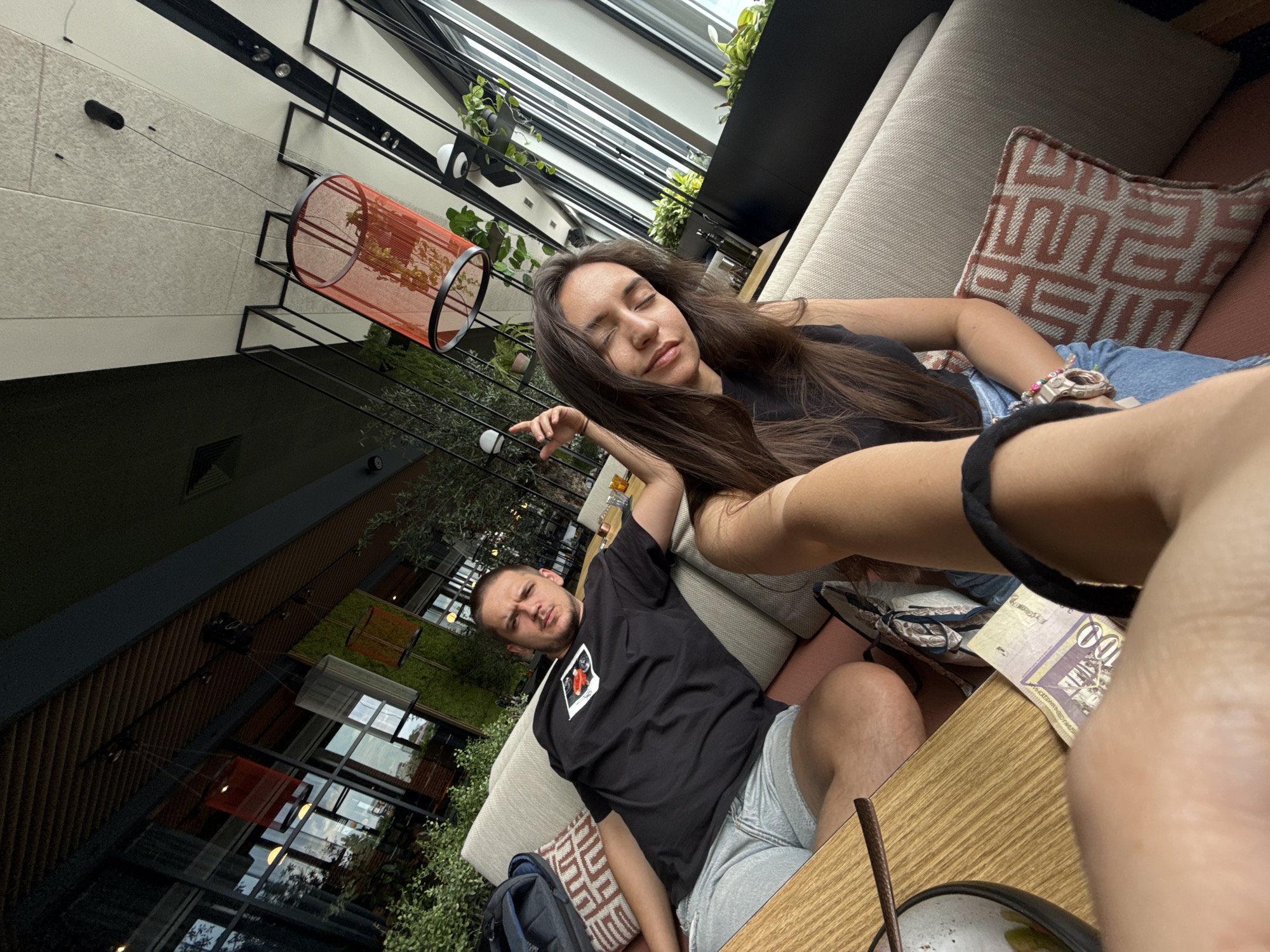A photo taken at a sharp angle shows a man and a woman in what appears to be a restaurant or lounge area in Plovdiv, Bulgaria. The woman is lying down with her eyes closed and long brown hair. She is wearing a black top and jeans. A man is sitting next to her, looking towards the camera with a slight frown. He is wearing a black t-shirt with a graphic and grey shorts.
They are sitting on a beige sofa with decorative pillows. On the table in front of them, there is money (likely Bulgarian Lev) and what appears to be a bowl of food. The environment suggests a modern architectural style with large windows and hanging planters. There are other architectural elements, including metal beams, a decorative orange cylinder suspended from the ceiling, and various plants, contributing to an upscale indoor-outdoor atmosphere.