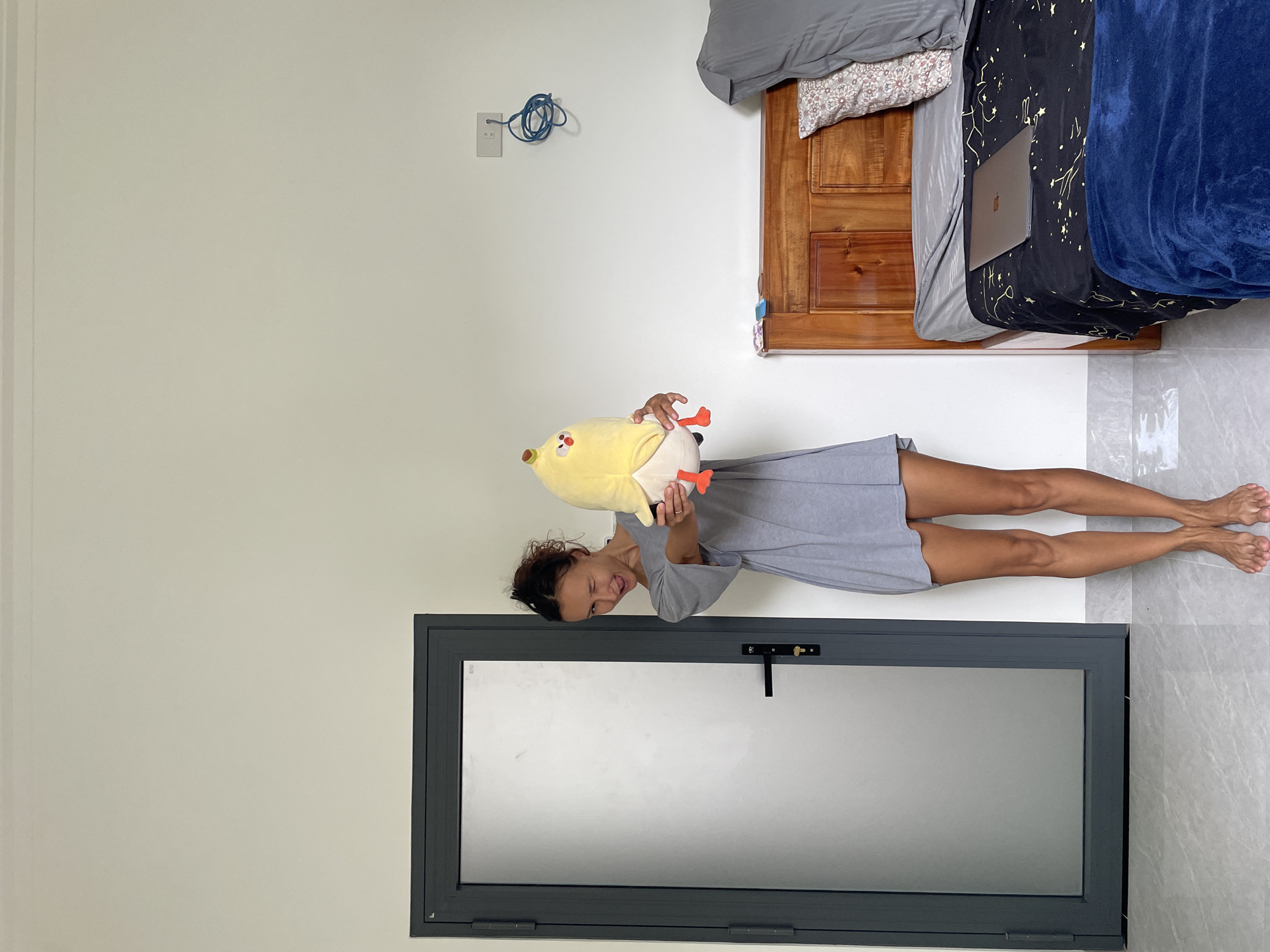 This image, originally rotated 90 degrees clockwise, captures a woman playfully posing indoors, likely in a bedroom or studio apartment in Mũi Né, Vietnam. The woman, appearing to be in her 20s or 30s with dark, curly hair tied back, smiles broadly while looking towards the camera. She is wearing a light grey, short-sleeved mini-dress and holds a large yellow plush toy resembling a chick or duck, complete with a small red beak and feet.
The room features plain white walls and a light grey tiled floor. In the background, a wooden bed frame is visible, dressed with a grey fitted sheet, a patterned pillow, and a striking black duvet adorned with yellow star and constellation patterns. A dark blue fuzzy blanket is draped over part of the bed, and a silver laptop is open on top of the duvet. On the wall above the bed, a white electrical outlet has a coiled blue cable hanging from it. Below the woman, a modern, dark grey framed window or door with frosted or reflective glass and a black handle extends across the lower portion of the frame. The scene conveys a cheerful and relaxed mood within a clean, bright, and contemporary living space. No discernible text is visible.