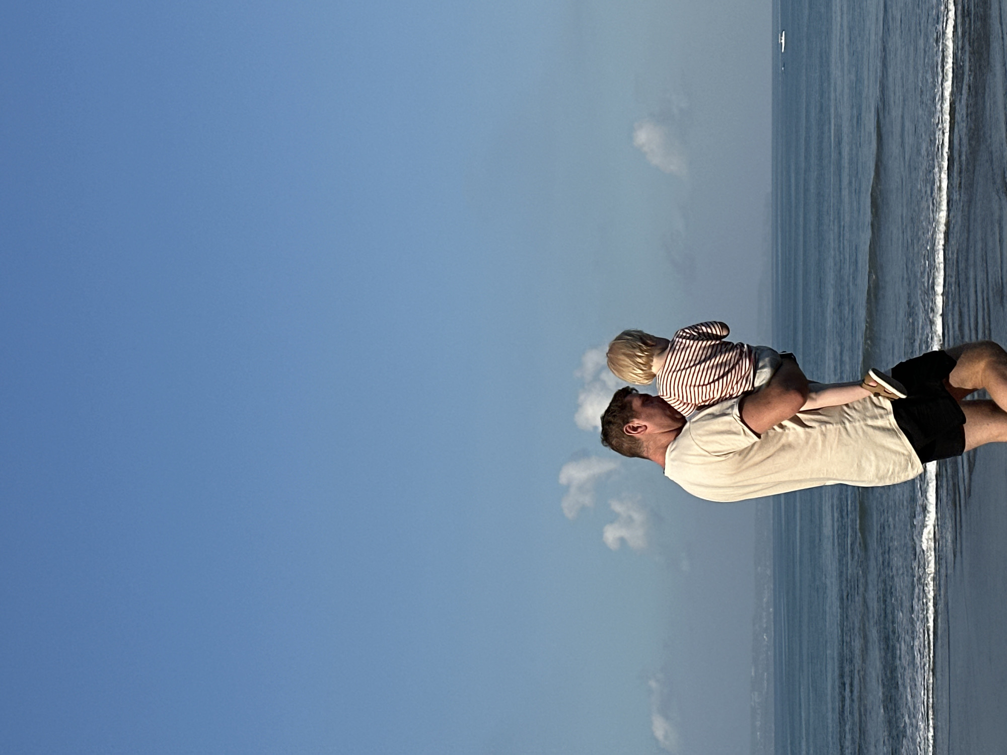 An adult male holding a young child is standing on a beach in North Kuta, Indonesia, facing the ocean. The male is wearing a light-colored t-shirt and black shorts, and the child is wearing a striped shirt. The ocean is calm with gentle waves, and there are a few clouds in the bright blue sky. In the distance, a small boat can be seen on the water. The scene appears peaceful and serene.