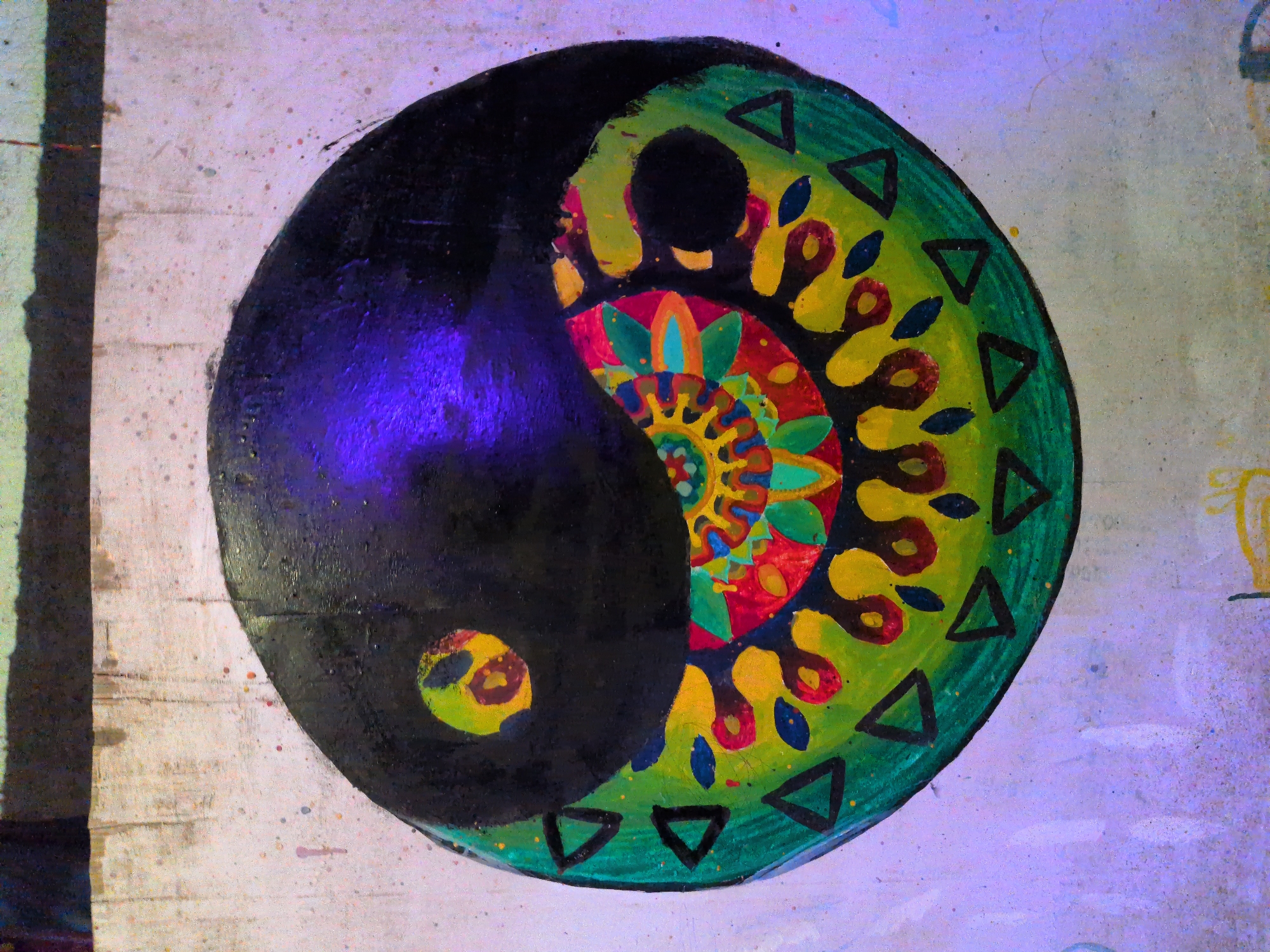 The image shows a close-up of a colorful Yin Yang symbol painted on a textured wall. The symbol is predominantly black, green, yellow, red, and blue, with intricate mandala-like patterns within the green and yellow sections. The black portion of the symbol has a reflective purple sheen, suggesting it might be painted with a glossy finish or illuminated by colored light. The background wall is a rough, off-white surface with visible paint splatters in various colors, indicating it's likely an outdoor or art space. The bottom edge of the image reveals a hint of a dark, horizontal band above the painted symbol, and further below, there are faint yellow markings on the wall that appear to be part of another artwork or graffiti.
This artwork is located on a wall in Leh, India, specifically in the area around the Hall of Fame, a museum dedicated to the history and heritage of the Indian Armed Forces in the region. The Hall of Fame is a popular tourist attraction in Leh.