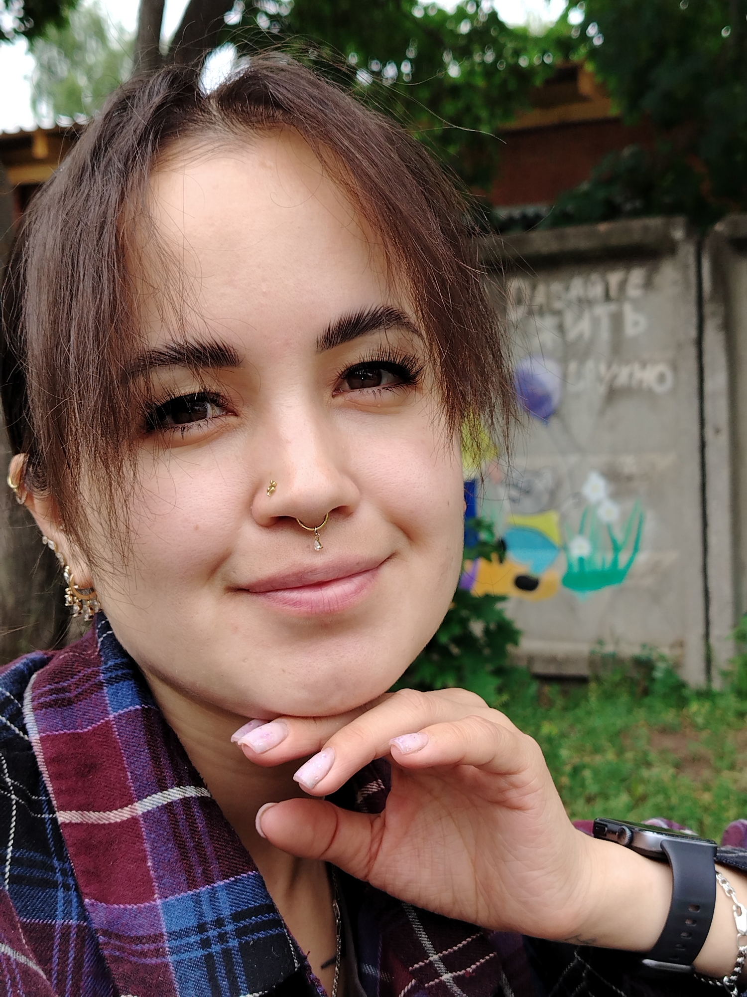The image is a close-up, front-facing portrait of a young woman with dark brown hair, styled with bangs. She has defined eyebrows, long eyelashes, and a nose ring with a small dangling charm. Her left ear is adorned with multiple gold earrings. She is wearing a purple and blue plaid shirt. Her right hand is resting on her chin, and her fingernails are painted a light pink. A dark smartwatch is visible on her left wrist.
The background is blurred but appears to be an outdoor urban setting. Behind the woman is a concrete wall with graffiti. To the left of the wall, trees and foliage are visible. The graffiti includes what appears to be text in Cyrillic letters and colorful drawings of abstract shapes and plants. The lighting suggests it is daytime, possibly overcast.
This scene is depicted in Noviy, Russia, an area known for its industrial buildings and residential districts, often characterized by concrete structures and pockets of greenery. The presence of graffiti on the wall suggests a public or semi-public space within the city. The overall atmosphere is casual and candid, likely taken outdoors.