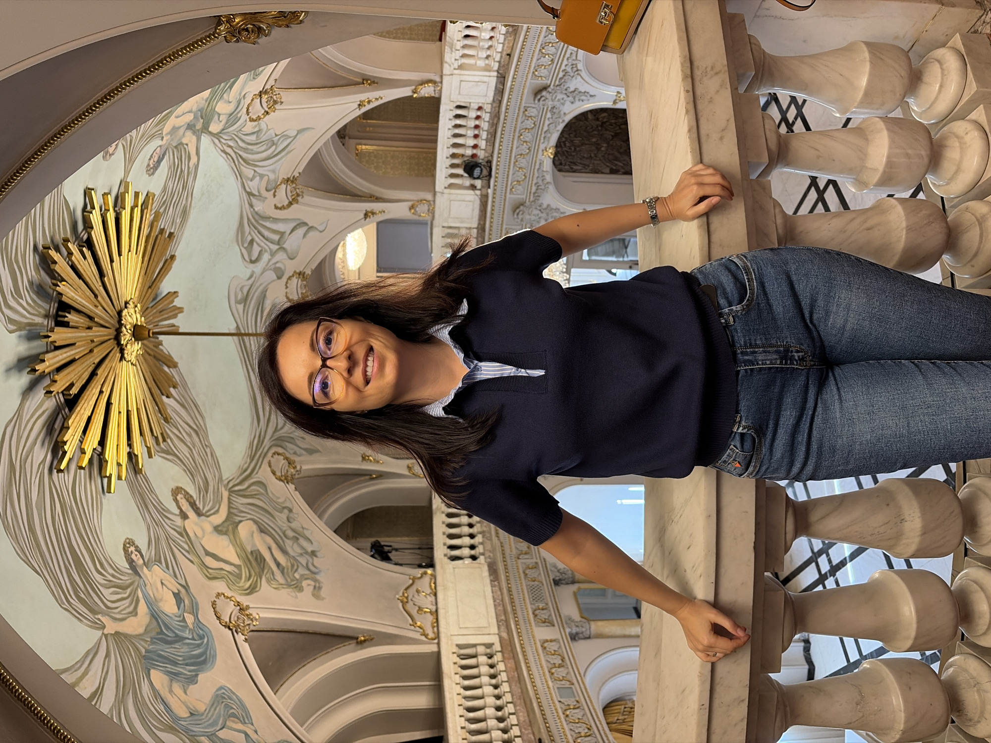 A woman stands leaning on a marble railing in a grand interior space, possibly a theater or opera house, with ornate balconies and ceiling frescoes. She has dark hair, wears glasses, a dark blue collared shirt, and blue jeans. A tan handbag is visible to her right. The architecture features classical columns, gilded decorations, and a large sunburst-like fixture on the ceiling. This location is identified as the National Palace of Culture in Sofia, Bulgaria.