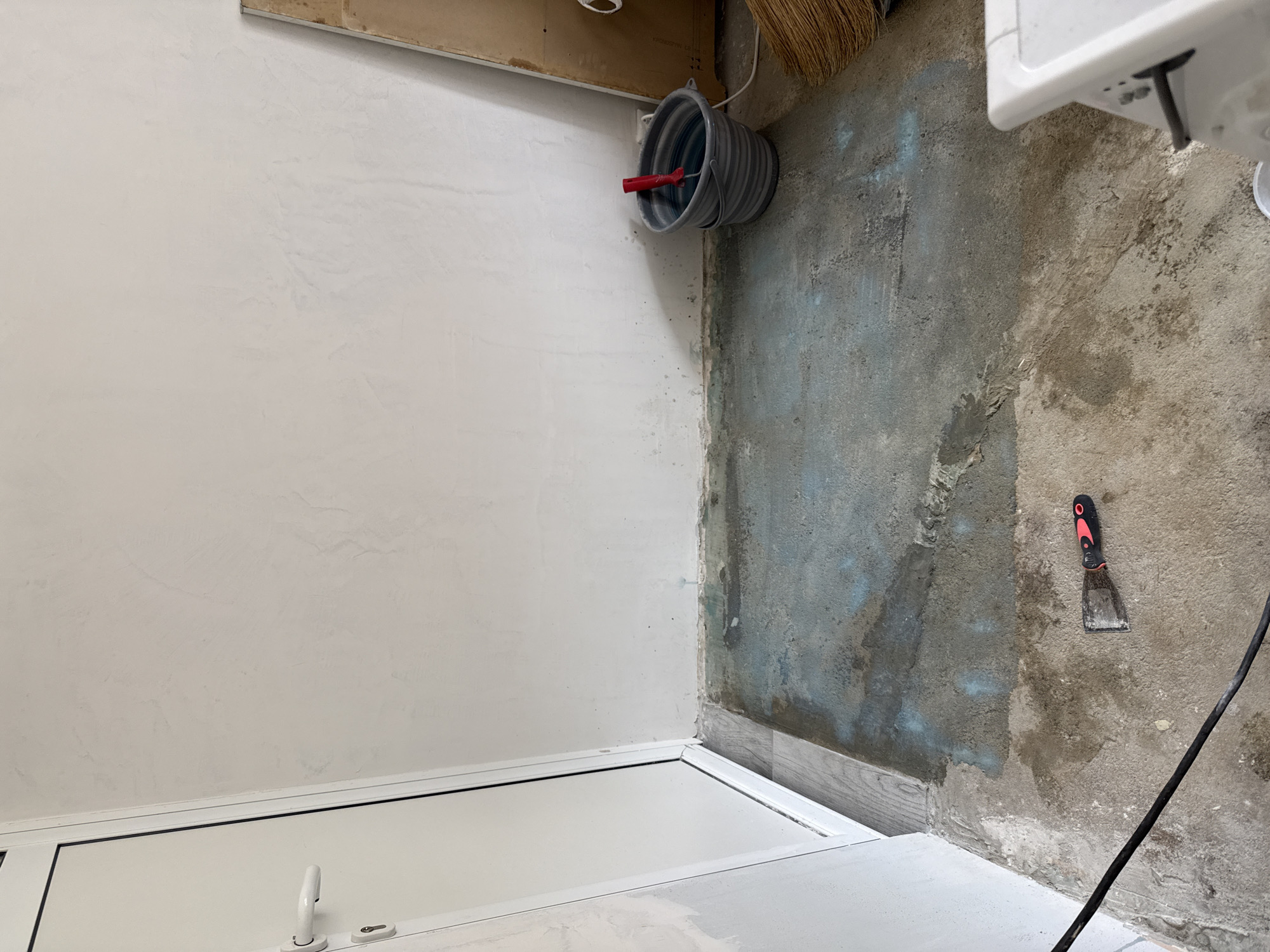 The image depicts an interior corner of a room, possibly under renovation. Walls are unfinished, showing areas of plaster and bare concrete. A closed white door is in the foreground. A gray bucket with a red-handled roller inside hangs on the upper right. A black cable runs diagonally across the lower right of the image. A scraper with a red and black handle is positioned on the bare concrete wall. The location appears to be a construction or renovation site, possibly in Stara Zagora, Bulgaria. No people are visible.