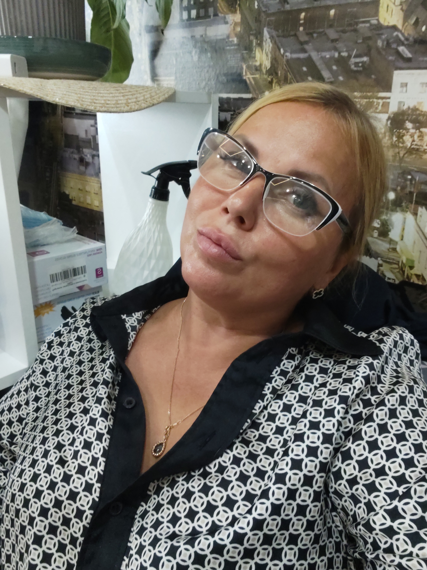 The image shows a close-up, medium shot of a woman with blonde hair and glasses, wearing a black and white patterned shirt. She is looking towards the camera with her head slightly tilted. Her left hand is resting on her chest. In the background, there are shelves with various items, including what appears to be a straw hat, a green bowl, and a white spray bottle. The wall behind the shelves has a wallpaper depicting a city scene. The lighting is bright, suggesting an indoor setting. The image does not provide any information about the location in the city of Dzerzhinsk, Russia.
