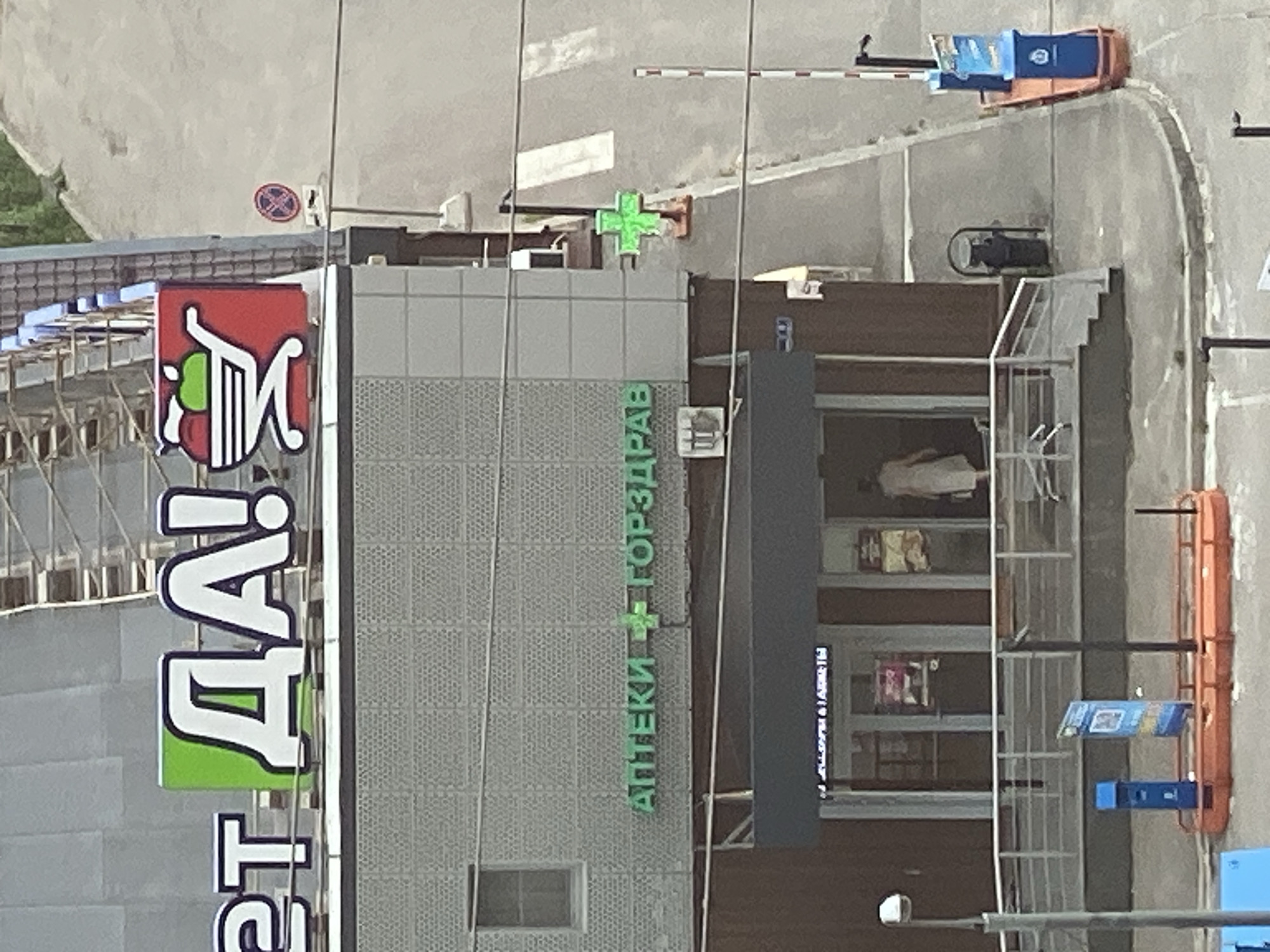 The image is an exterior, medium-distance, elevated shot of a commercial building. The building has a modern facade with sections of grey textured panels and a darker brown lower level. A prominent sign on the upper facade features large white letters spelling "ДА!" with a green background element and a red shopping cart graphic containing an apple. Below this, a green sign reads "АПТЕКИ + ГОРЗДРАВ" with a green cross symbol. A separate green cross sign is mounted on a pole on the right side of the building. The entrance to the building is visible with double glass doors and a ramp leading up to it, with a person in a light-colored dress walking inside. To the left of the entrance, there are blue and yellow informational signs on stands. A parking barrier and two blue trash receptacles are visible on the right side of the image. The ground area suggests a paved street or parking lot. The overall scene is static, with no evident actions or interactions beyond the person entering the building. The location appears to be a commercial area in a city, possibly on a sunny day given the lighting and shadows.