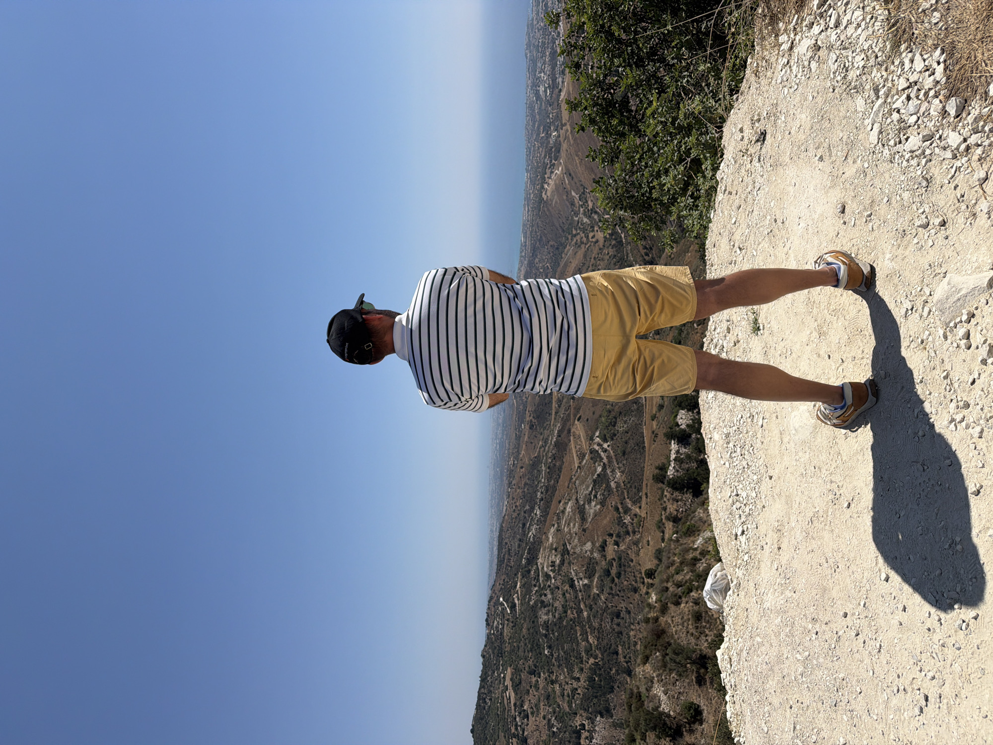 A man stands on a dirt path overlooking a coastal city with the sea in the background. He is wearing a striped polo shirt, yellow shorts, a baseball cap, and sneakers. The landscape is dry and hilly, with sparse vegetation. The sky is clear and blue. The man is facing away from the camera, with his arms crossed. The scene is set in Peyia, Cyprus, on a bright, sunny day.