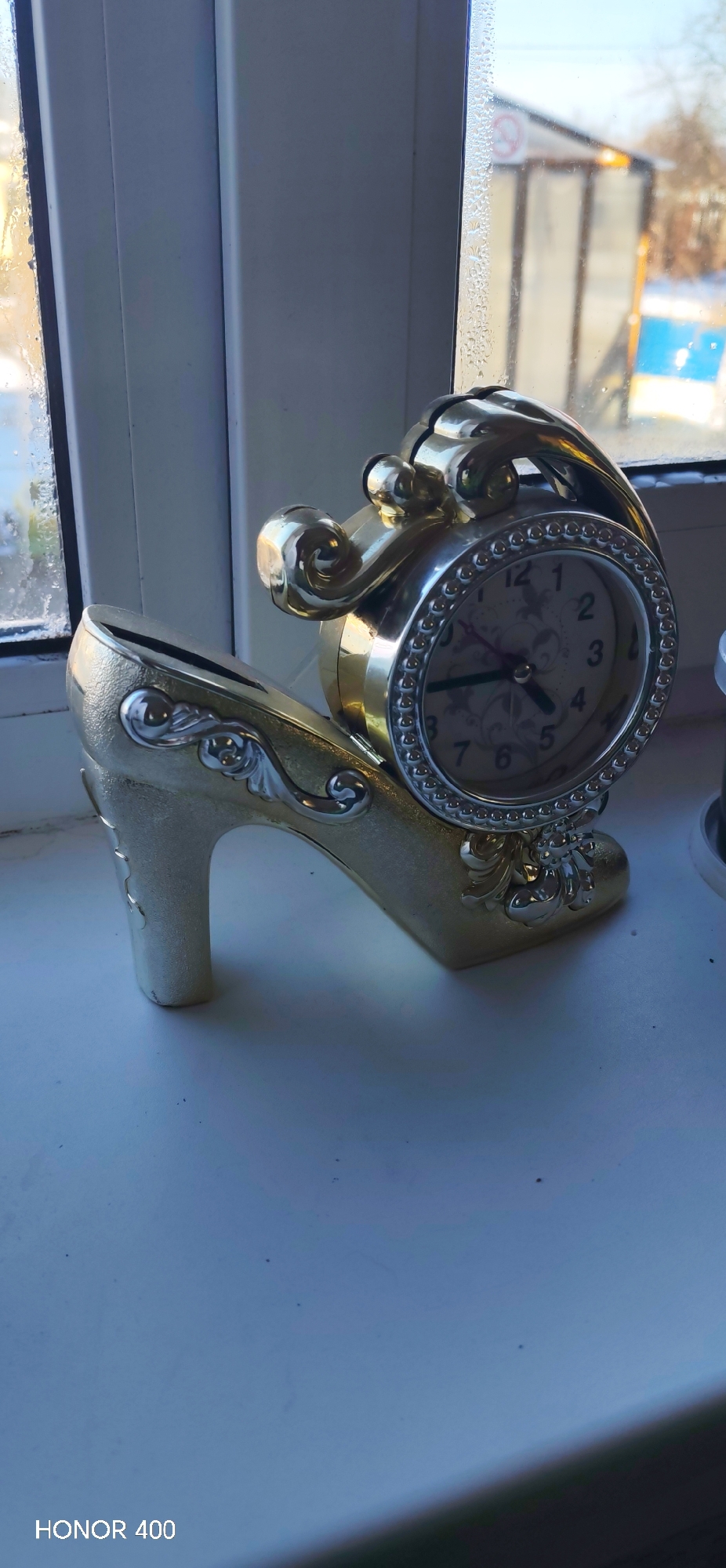 A clock in the shape of a high-heeled shoe sits on a white windowsill. The clock face shows a time of approximately 5:54. The view outside the window shows a building or structure. The image was likely taken with a Honor 400 camera. The location is specified in the prompt, but I cannot verify it from visual information.