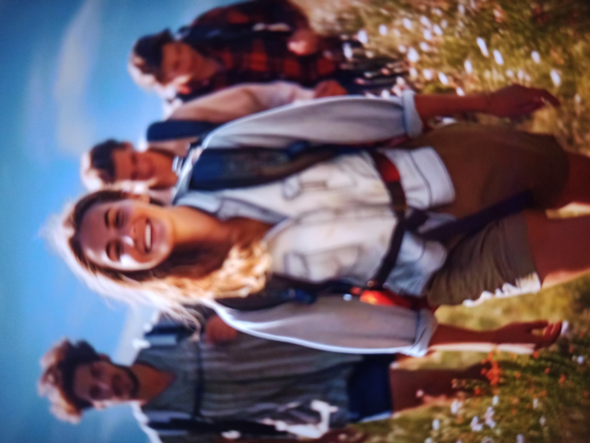 Four individuals are hiking in a rural outdoor environment under a bright blue sky. The person in the foreground is a young woman with blonde hair, smiling and looking towards the camera. She is wearing a light-colored button-down shirt with rolled-up sleeves, khaki shorts, and a backpack. Behind her, three other people are also hiking, all wearing backpacks. The person immediately behind her appears to be a young man with dark, curly hair. Further behind them, two more individuals are visible, one wearing a plaid shirt. The environment is characterized by grassy terrain with scattered wildflowers, suggesting a natural, undeveloped area. There is no indication of the city of Balakliya, Ukraine, within the image.