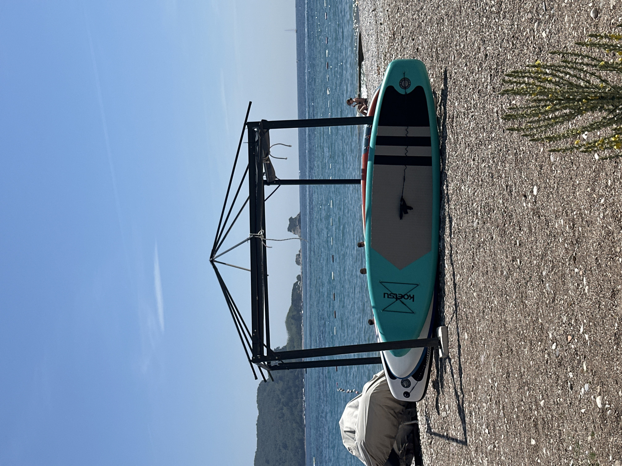 The image displays a sunny coastal scene, likely a pebble beach in Budva, Montenegro. In the foreground, two paddleboards are resting on the pebbled ground. The most prominent paddleboard is teal, grey, and black, with the brand name "KOETSU" visible on its teal section, along with a black elastic cord system on the deck pad. A second, mostly white paddleboard with red and grey accents is partially visible beside it.
Behind the paddleboards, a dark metal rack or frame structure stands, possibly used for drying or storing water sports equipment. A boat or jet ski, covered with a light-colored tarp, is parked behind this structure. The calm, blue sea stretches into the middle ground, where a lone person is visible sitting or crouching near the water's edge. Distant landmasses, appearing as hills or mountains, line the horizon across the water.
The sky above is clear and bright blue, suggesting a beautiful, sunny day. To the far right, a small portion of a green bush adorned with tiny yellow flowers adds a touch of natural detail. The overall scene suggests a relaxed atmosphere conducive to beach activities and water sports.