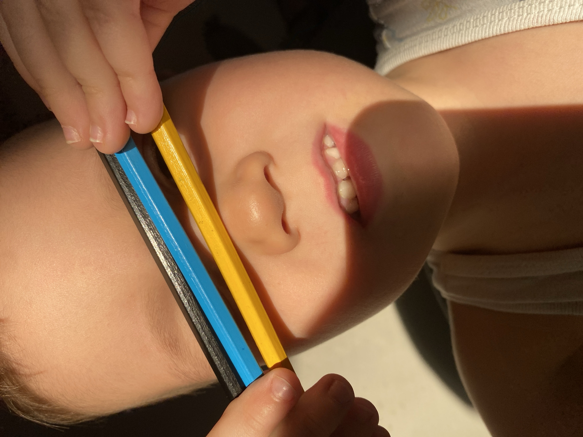 A close-up, eye-level shot depicts a child lying down, with three colored pencils (black, blue, and yellow) held across their face, near the eyes. The child's mouth is slightly open, revealing a few teeth. One ear is visible. The person holding the pencils is partially visible. The skin tone is fair. Light and shadows play across the scene. This image was not taken in Chaikovsky, Russia.