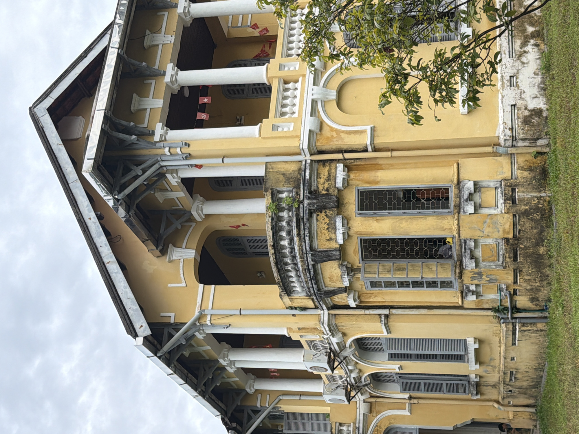 The image shows a large, two-story yellow building with white trim and ornate architectural details. The building has a covered balcony supported by white columns on the upper floor, and arched windows with grey shutters on both levels. Red flags are visible hanging from the balcony. The exterior of the building appears weathered, with some peeling paint and staining on the lower portion. An air conditioning unit is mounted on the left side of the building. Green grass covers the ground in front of the building, and a tree with green leaves is partially visible on the right. The sky is overcast.
