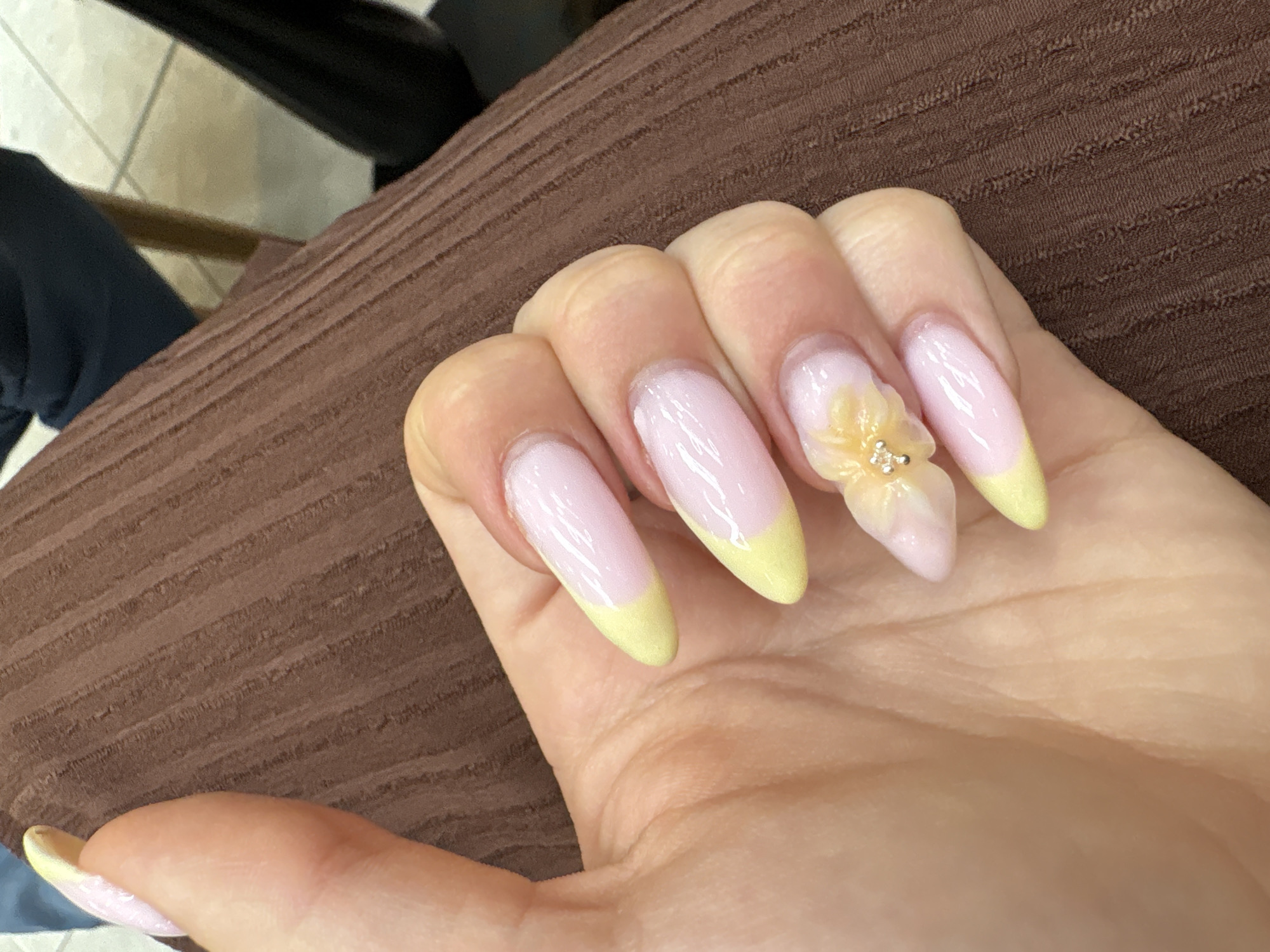 The image shows a close-up of a human hand with long, almond-shaped nails. The nails are painted with a light pink base and have a yellow French tip. One nail is adorned with a 3D flower. The hand is resting on a textured brown surface. The background includes a black chair leg and a glimpse of a tile floor. No information in the image suggests that the photo was taken in Burgas, Bulgaria.