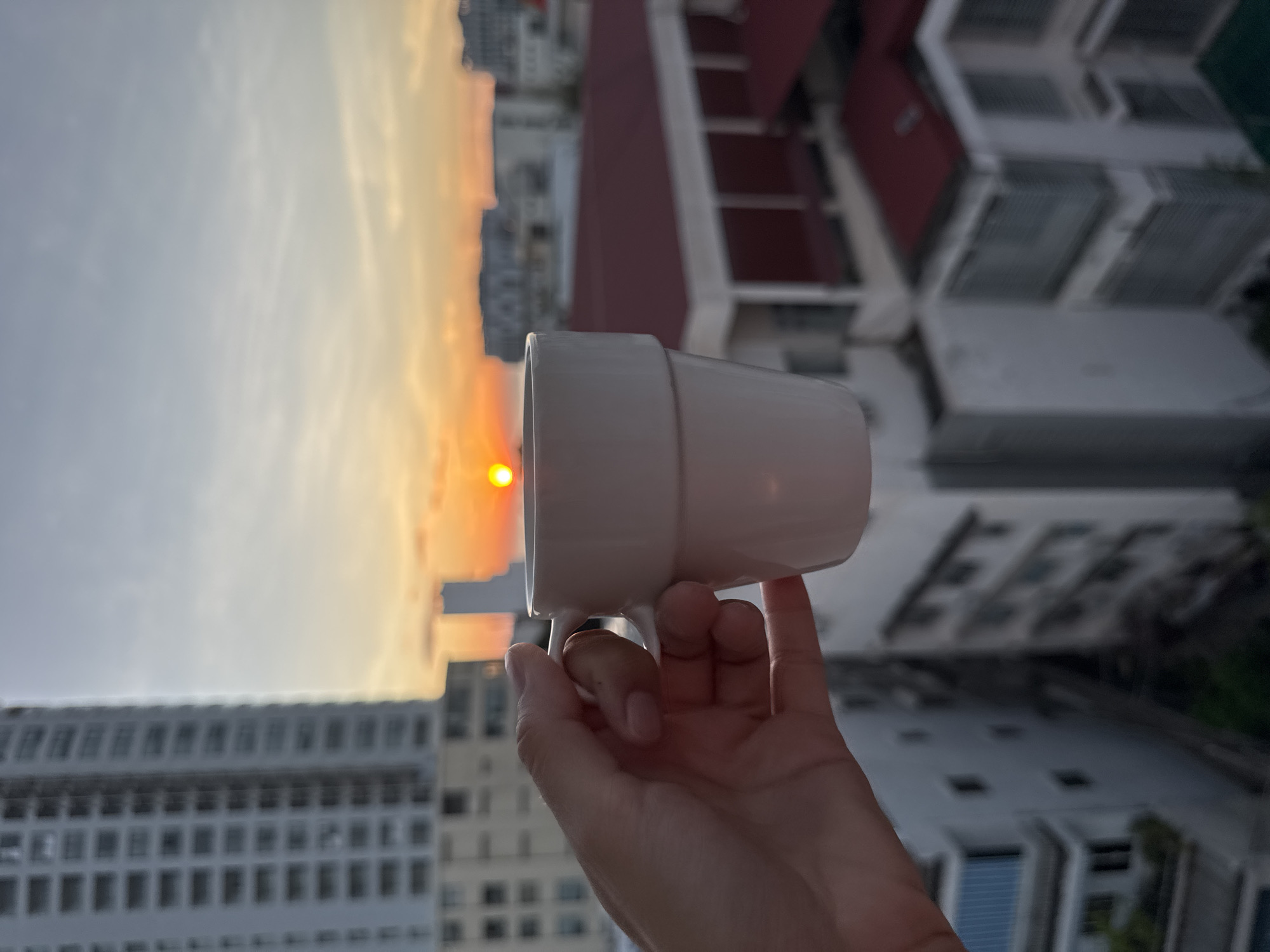 A hand holds a white mug in the foreground against a city skyline at sunset. The mug has a simple, modern design with two horizontal bands. Behind the mug, the sun is visible as a bright orange orb low on the horizon, casting a warm glow across the sky. The sky transitions from a bright orange near the sun to a pale yellow and then a soft blue. In the mid-ground, buildings of varying heights are visible. To the left, a tall, modern apartment building with many windows dominates the frame. To the right, lower buildings with red tiled roofs are prominent. The scene appears to be from a vantage point overlooking the city, likely from a hotel room or apartment balcony in Nha Trang, Vietnam, given the architectural style and coastal city feel. The overall mood is peaceful and serene, capturing a moment of quiet contemplation with a view.