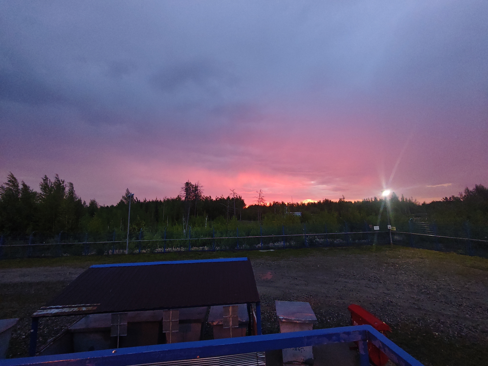 The image captures an outdoor scene at dusk, dominated by a vibrant, colorful sunset. The sky transitions from a deep blue-purple at the top to striking shades of pink and magenta closer to the horizon, where the sun's light is most intense, creating a radiant glow. Some clouds are scattered across the sky, catching these brilliant colors.
Across the mid-ground, a dense line of dark evergreen trees forms a silhouette against the bright horizon, with a few bare, stark trees visible. In front of the forest, a chain-link fence stretches horizontally, marking a boundary. A tall pole with a bright, artificial light source stands on the right side, illuminating a small area and casting a visible light ray.
The foreground appears to be a cleared, gravel or dirt area. The viewpoint is from a slightly elevated position, indicated by a prominent blue railing at the very bottom of the frame. In the lower-left, a dark, low-slung structure with a blue trim, possibly a shed or industrial equipment housing, features multiple compartments. Another dark, cuboid structure is partially visible to its right. A red box or cabinet, possibly a utility or safety box, is seen in the bottom right corner.
No specific human activity is visible, suggesting a tranquil moment at day's end. The overall impression is one of dramatic natural beauty contrasting with utilitarian man-made structures. There is no legible text visible on any objects or signs within the image.