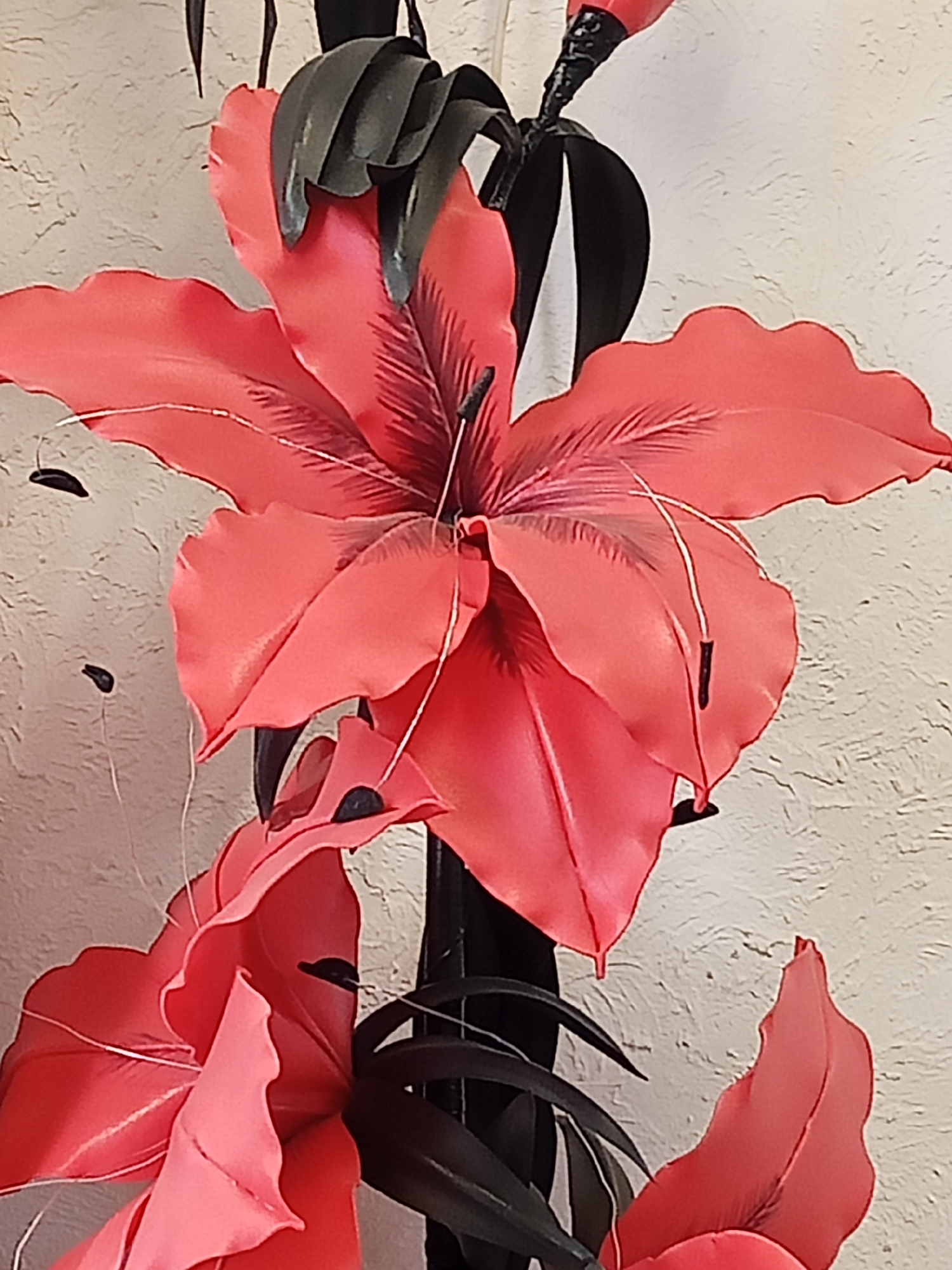 The image shows a close-up of artificial flowers, likely made of fabric or plastic. The dominant color is a vibrant coral pink, with darker tones defining the petal structures and veins. Several large petals are visible, some unfurling outwards, while others are more tightly closed. Thin, white stamen-like filaments tipped with black can be seen extending from the center of some flowers. The flowers are attached to a dark stem or stalk, which is surrounded by dark green or black leaf-like structures. The background is a textured, light-colored wall, possibly painted or wallpapered. There are no people or animals in the image. The scene appears to be indoors, given the presence of the wall as a backdrop and the artificial nature of the flowers. There is no information in the image to indicate its location within Panino, Russia.