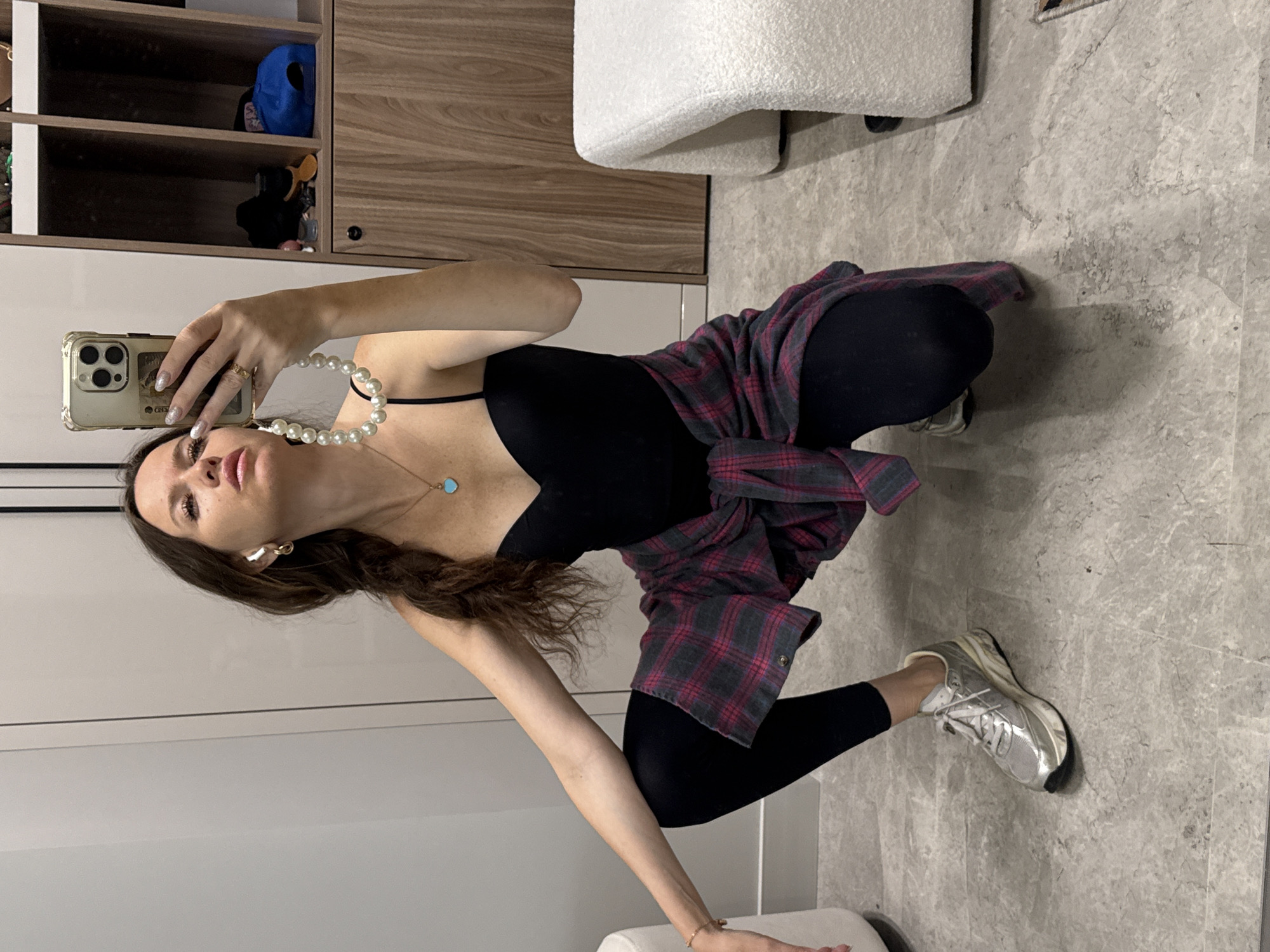 A young adult woman is crouched down, taking a selfie with her smartphone. She has long brown hair tied in a braid, wears a black leotard, black leggings, a plaid shirt tied around her waist, and silver sneakers. She has white earbuds in her ears and is wearing a pearl necklace with a turquoise heart pendant. The background appears to be a dressing room or changing area, with a mirrored wall, wooden shelving units displaying various items, and a textured white bench. The floor is tiled with a grey, marble-like pattern. The lighting is artificial and bright. There is no indication of the location being in Khimki, Russia.