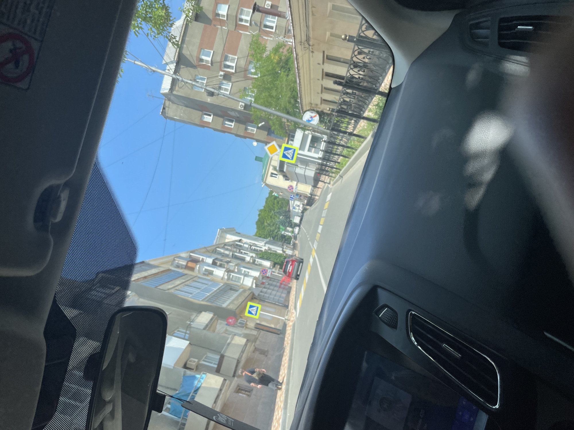 The image is taken from the interior of a car, looking out through the windshield. The car is driving on a street in the city of Stavropol, Russia. On the left side of the street are multi-story buildings with balconies, and a sidewalk where a man in dark clothing is walking with a bag over his shoulder. A red car is visible driving ahead in the same direction as the camera car. On the right side of the street are more buildings, including a taller building with red and gray horizontal stripes, and a lower beige building with an ornate metal fence in front of it. Several traffic signs are visible, including a pedestrian crossing sign and other regulatory signs. The sky is clear and blue, indicating a sunny day. The interior of the car shows the dashboard, rearview mirror, and part of the windshield with a mesh pattern along the top edge.