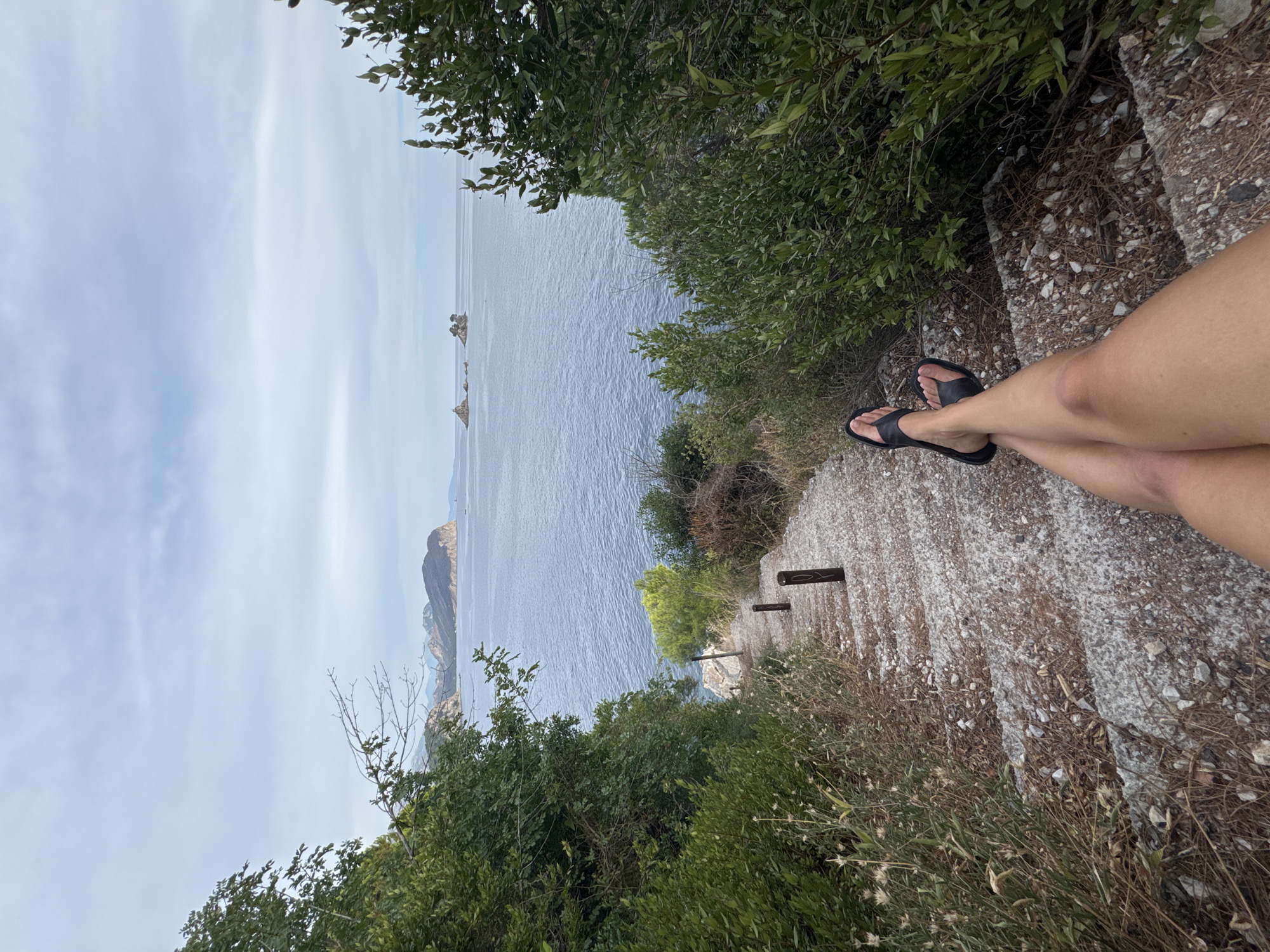 A person's legs are visible, crossed and wearing black sandals, as they sit on a stone staircase overlooking the sea. The scene is set in Mirište, Montenegro, with lush green foliage framing the view. The sea is a deep blue, with distant rocky outcrops emerging from the water. The sky is mostly clear with some scattered clouds. The time of day appears to be late morning or early afternoon, with bright, diffused sunlight. The person is seated on a weathered stone staircase, with dry grasses and small rocks scattered around. Two dark wooden posts are visible along the staircase, possibly for support or as markers. The overall impression is one of peaceful relaxation and appreciation of the natural beauty of the location.