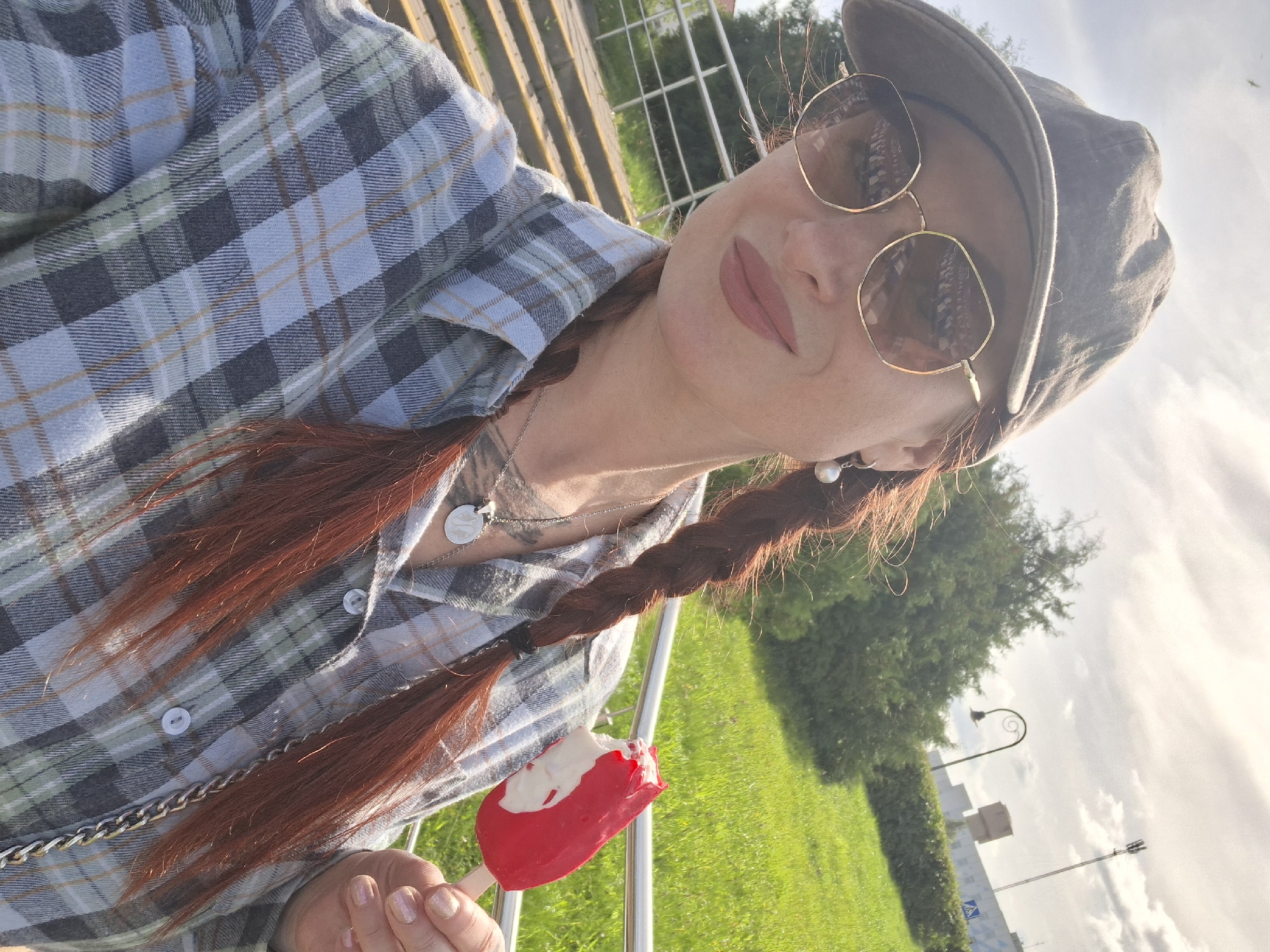 A woman with auburn hair, styled in two braids, is photographed outdoors. She wears a grey and blue plaid shirt, a grey baseball cap, and hexagonal sunglasses. She is holding a partially eaten red and white popsicle. In the background, there is a grassy area, trees, stairs, a metal railing, a streetlamp, and a cell tower. The lighting suggests it is daytime, likely with the sun partially obscured by clouds. The image is taken from a selfie perspective. The setting appears to be a park or public outdoor area within the city of Myadzyel, Belarus, indicated by the presence of urban infrastructure and manicured green spaces.