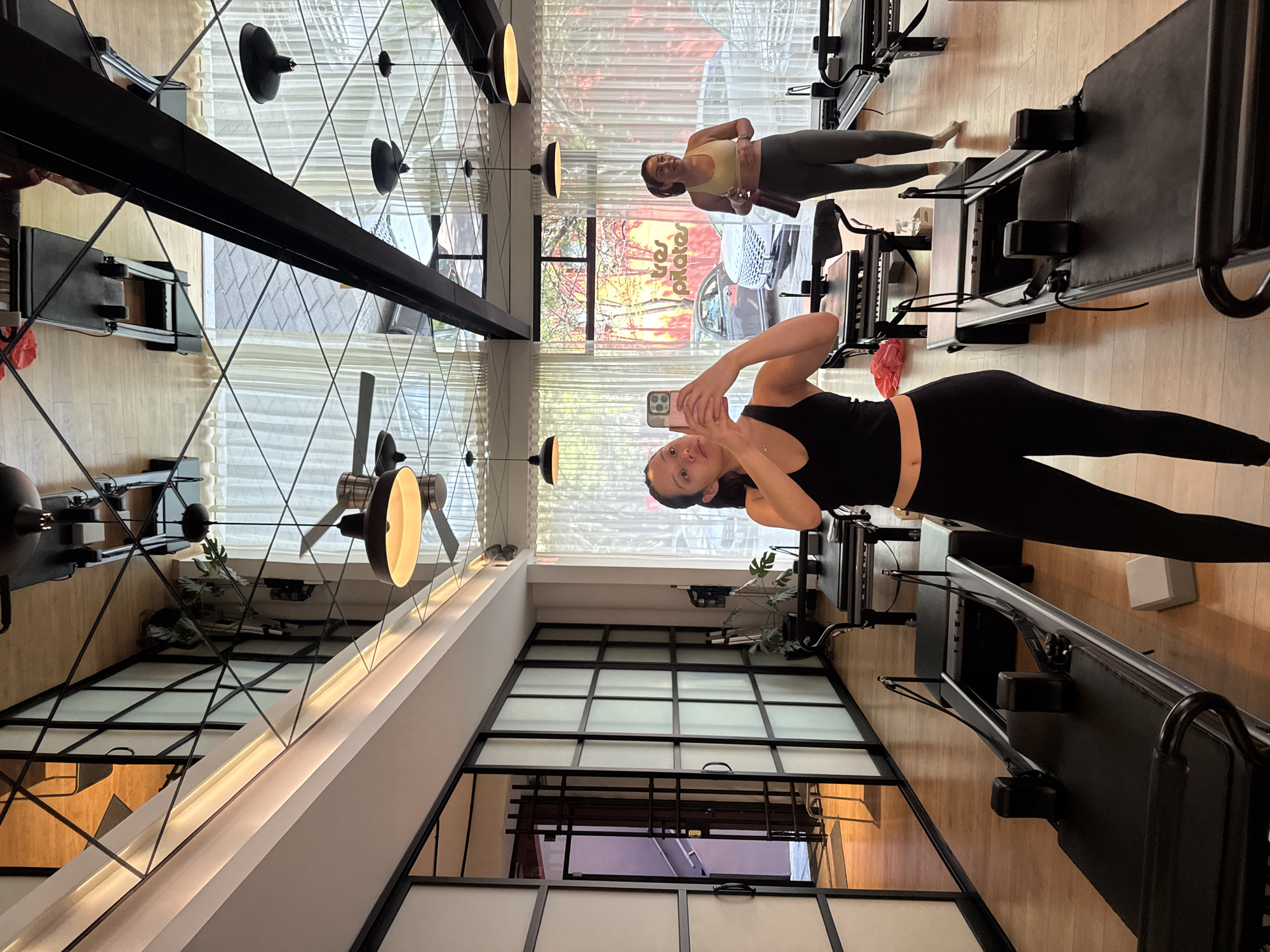 The image captures a vibrant, well-lit Pilates studio, likely in Puebla, Mexico, during the daytime. Two women are present, both dressed in athletic wear.
In the foreground on the right side of the frame, a woman wearing a black tank top and black leggings is taking a selfie with a light-colored smartphone. She holds the phone with both hands, looking directly at the camera with a slight smile.
In the mid-ground, towards the center-left, another woman stands near a black Pilates reformer. She is dressed in a light yellow sports bra and dark grey leggings and holds a dark water bottle in her hand. Her gaze is directed away from the camera, towards the large windows that line the back wall of the studio.
The studio itself features light wooden flooring and multiple black Pilates reformers. The back wall is dominated by expansive windows covered with sheer white curtains, through which natural light floods the space. Beyond the windows, hints of greenery and what appears to be an outdoor urban scene are visible. Reflected in the window is partial text that reads "tives pilates studio," indicating the name of the establishment.
The left wall of the studio features a large mirror, reflecting parts of the ceiling and opposite walls. The ceiling has a distinctive crisscross pattern of dark lines, along with modern pendant light fixtures and ceiling fans with integrated lights. The overall impression is one of a clean, modern, and inviting fitness environment.