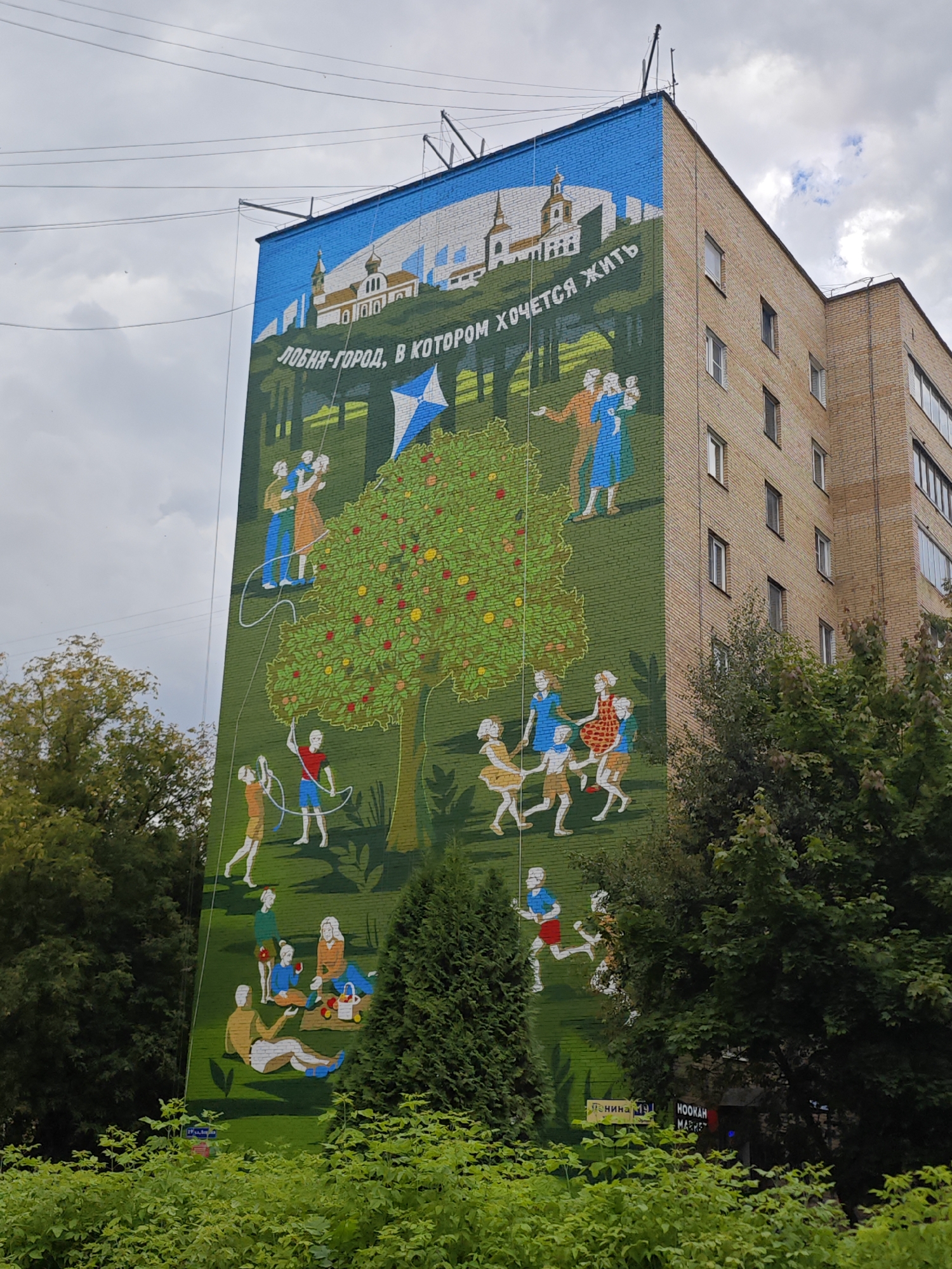 The image depicts a vibrant mural painted on the side of a multi-story brick building in Lobnya, Russia. The mural, taking up the majority of the building's facade, features a pastoral scene with stylized figures engaged in various outdoor activities. Dominating the center is a large, green, fruit-laden tree, with red and yellow fruits. Around the tree, people, including adults and children, are shown flying a kite, dancing, playing, and having a picnic. The upper section of the mural depicts a cityscape with white buildings, possibly churches, and a blue sky. Text in Russian, reading "ЛОБНЯ-ГОРОД, В КОТОРОМ ХОЧЕТСЯ ЖИТЬ" (Lobnya - a city where one wants to live), is prominently displayed across the mural. The building itself is a typical Soviet-era apartment block with visible windows. Lush green trees and foliage partially obscure the lower portion of the building and the mural. In the lower right, a sign for "HOOKAH MARKET" is visible. The sky is cloudy, suggesting overcast weather.