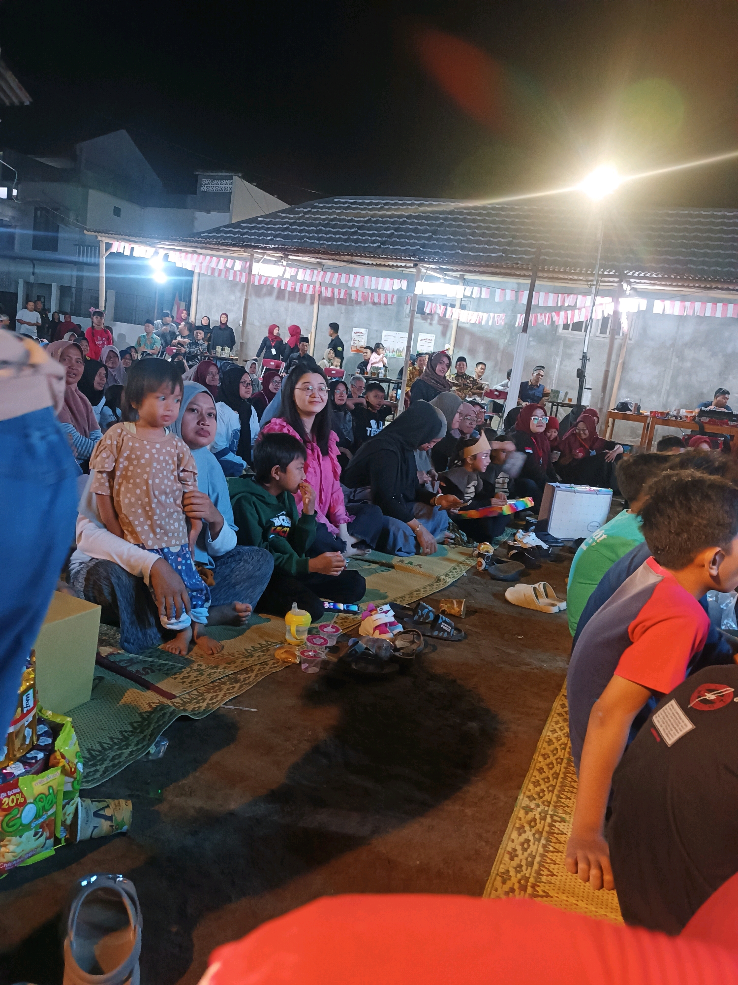 The image depicts an outdoor gathering at night, likely a community event, in Jetis, Indonesia. A group of people, including adults and children, are seated on mats on the ground, facing away from the camera. They are illuminated by overhead lights and decorative string lights adorned with Indonesian flags. The background features a covered structure with a tiled roof and a building. Some individuals are holding snacks and drinks, suggesting a festive or celebratory atmosphere. The ground is a mix of dirt and mats, with some scattered footwear and bags. The overall scene conveys a sense of communal activity and social gathering in a local setting.