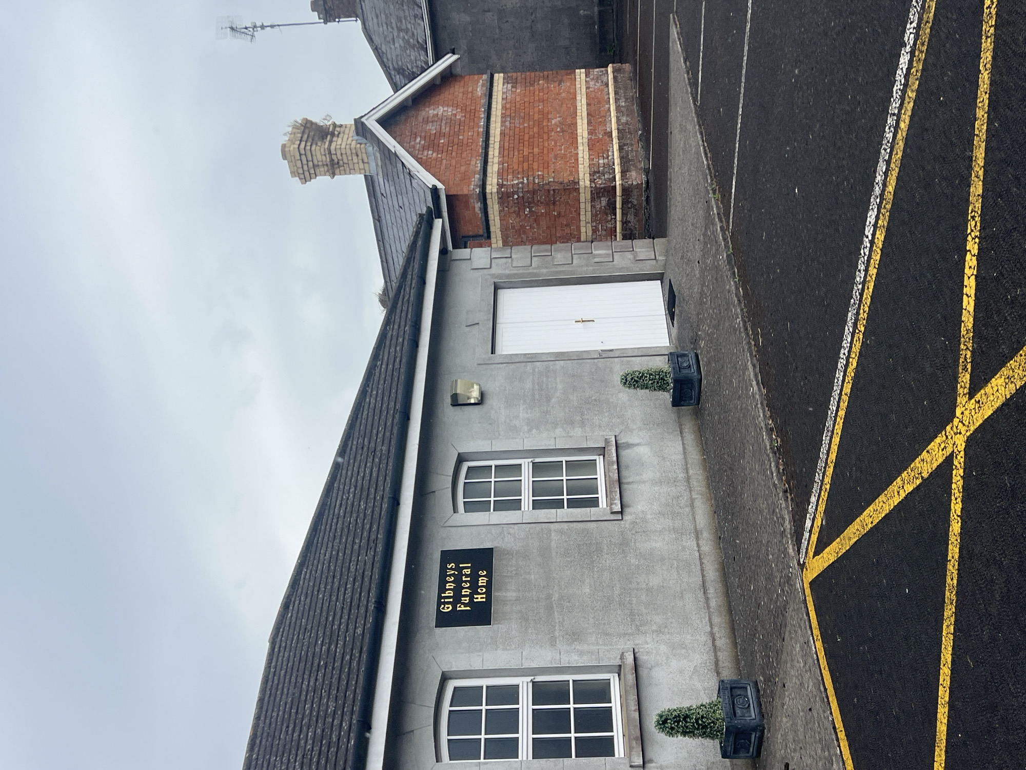 A grey, single-story building with a dark roof and two white-paned windows is visible. A black sign with gold lettering that reads "Gibneys Funeral Home" is mounted on the wall between the windows. To the right of the grey building, a smaller, two-story structure made of red brick with decorative horizontal yellow bands is attached. This brick structure has a peaked roof and a chimney. A white double door is visible on the left side of the grey building. Two square planters with small green conical bushes are positioned on the pavement in front of the grey building. The scene is outdoors, with a paved area in the foreground marked with yellow and white parking lines. The sky is overcast and grey. There are no people visible in the image.