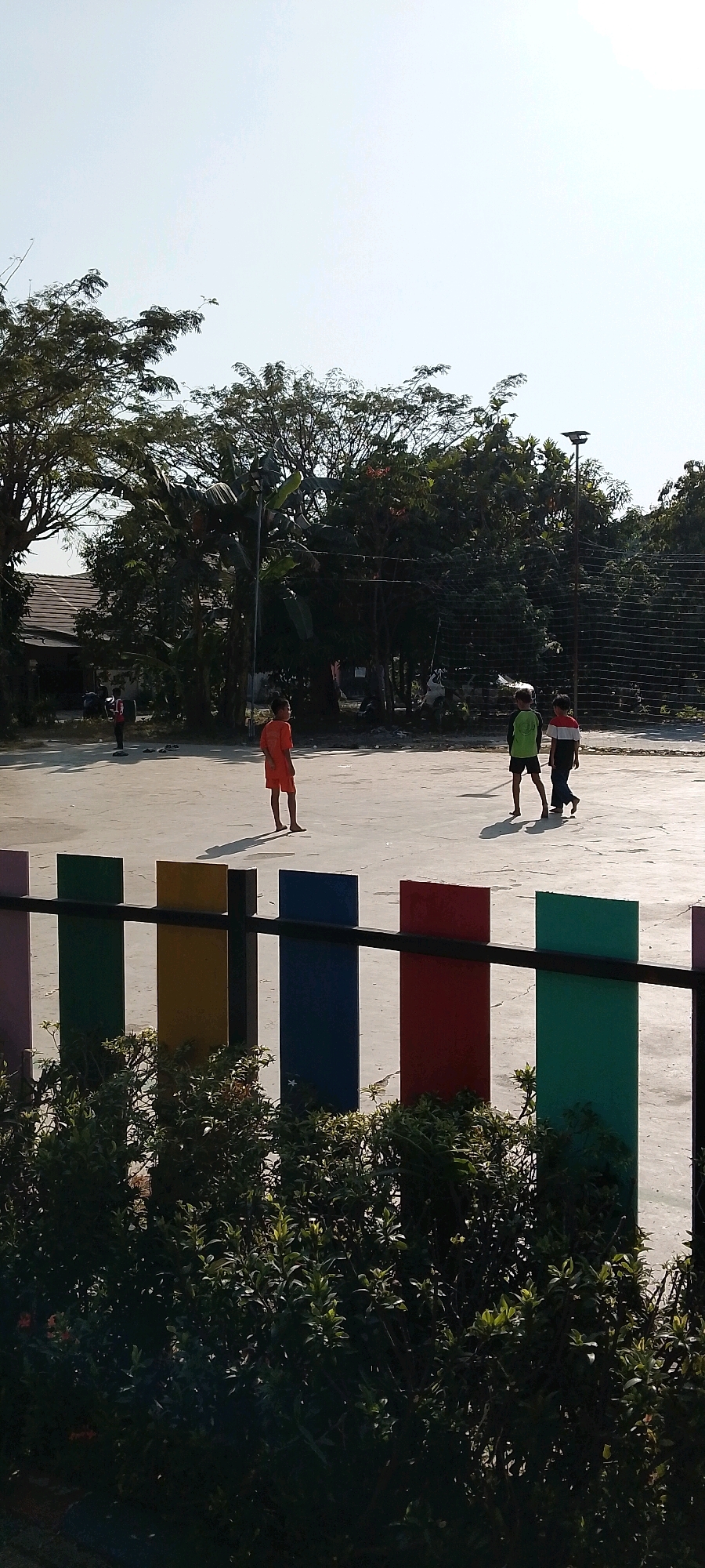 The image depicts an outdoor scene in Satriamekar, Indonesia, on a bright, clear day with a pale sky. In the foreground, a low-lying hedge obscures the bottom of a fence composed of vertical posts in alternating colors: pink, green, yellow, blue, red, and green. Beyond the fence, a paved court area is visible, casting long shadows from the sun. Three young boys are present on the court. One boy, wearing an orange outfit, stands facing away from the camera, looking towards the left. Two other boys are walking away from the viewer towards the right; one is wearing a green and black shirt and shorts, while the other is wearing a striped shirt and dark pants. In the background, dense foliage from various trees dominates the midground, with some leaves appearing tropical, like those of a banana tree. A building with a tiled roof is partially visible behind the trees to the left. Further back, a net, possibly for a sports court, stretches across the scene, and a lamppost is visible near the right edge of the frame. The overall impression is of a recreational or park area.
