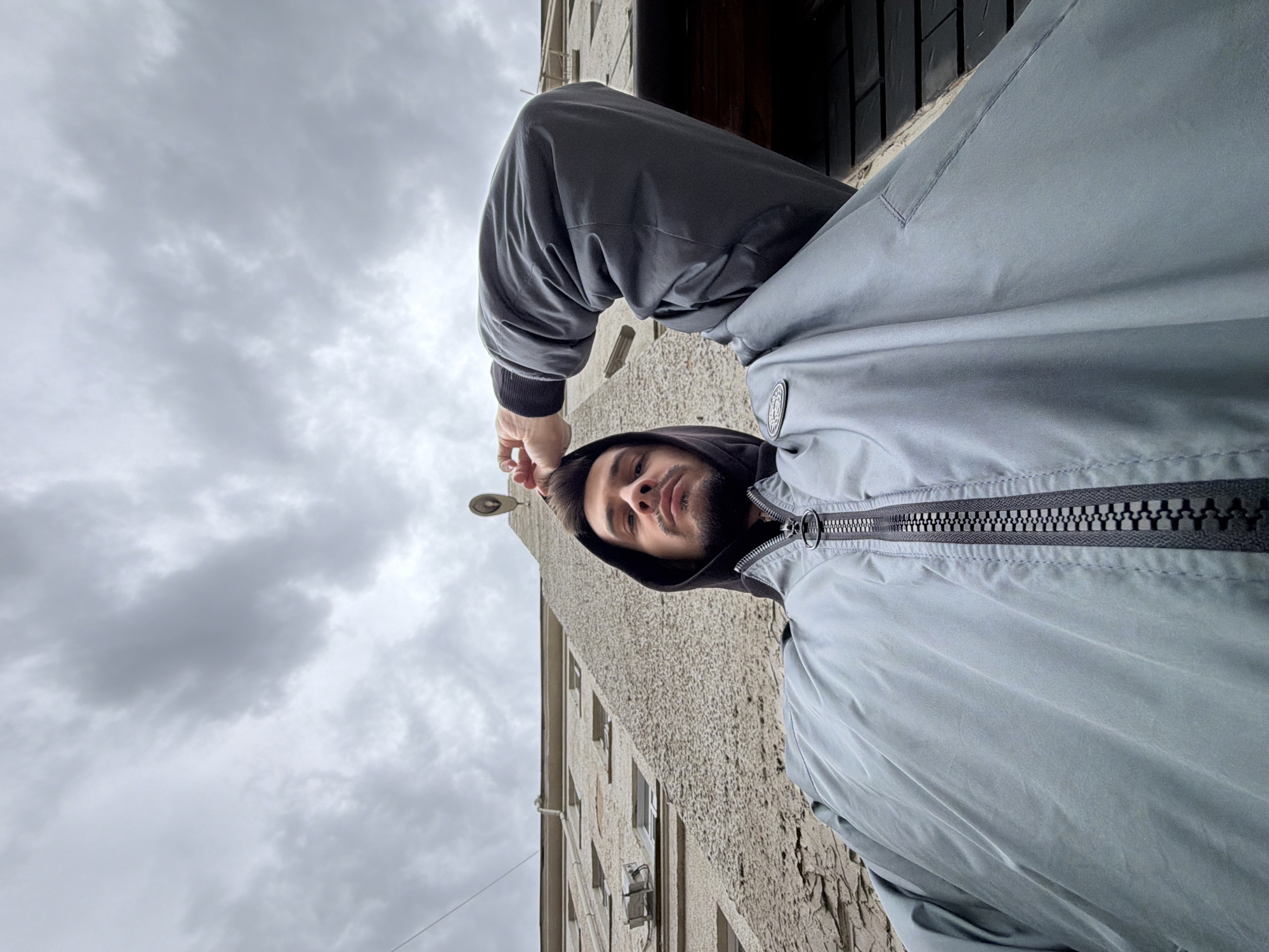 A low-angle, medium shot depicts a man with brown hair and a beard wearing a black hoodie underneath a zippered gray jacket. He's standing near a brick building with visible windows. Overcast skies are in the upper left. The location is in the city of Krasnoyarsk, Russia. A street lamp is visible above the man's head.
