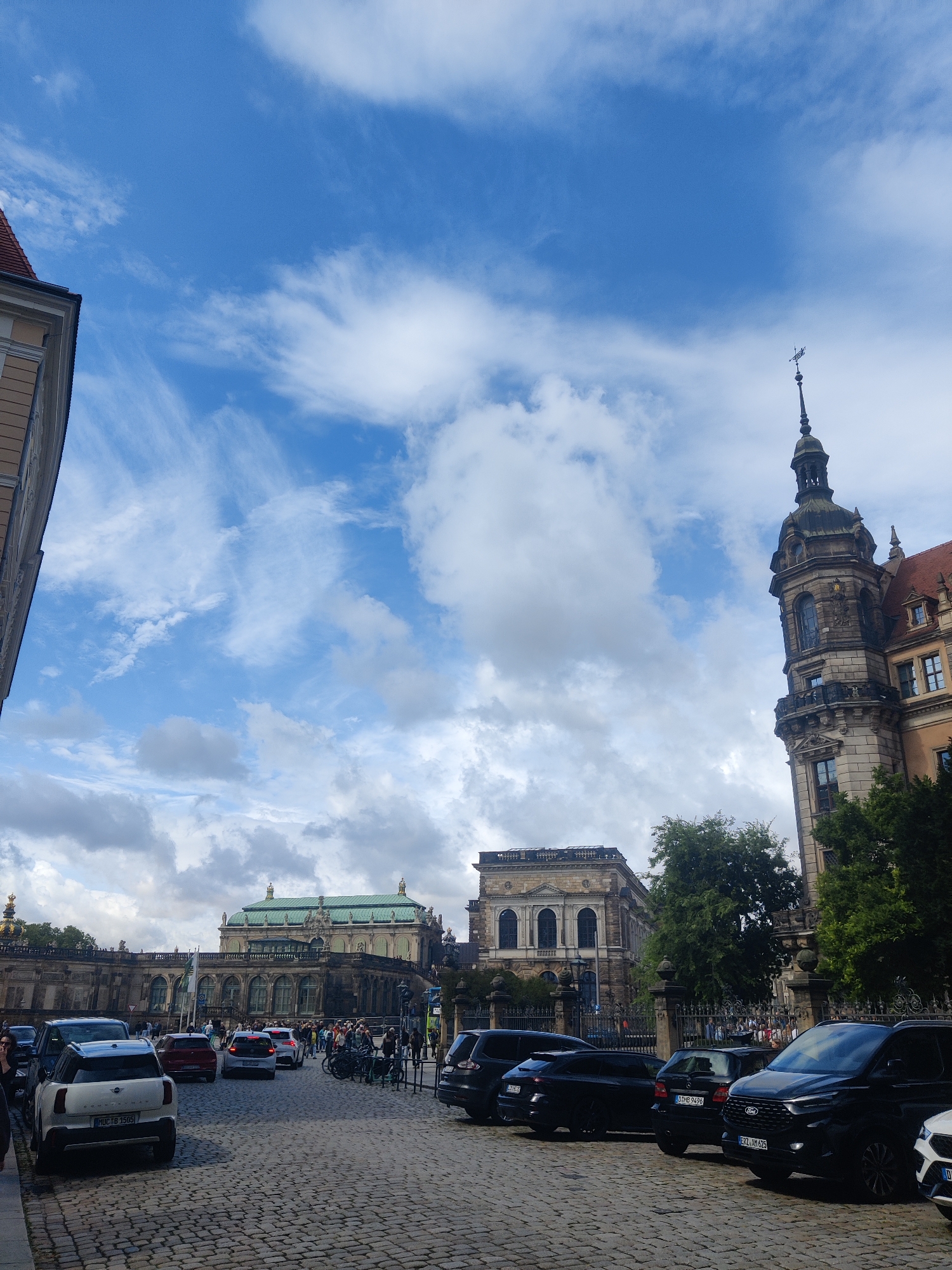The image shows a view of a cobblestone street in Dresden, Germany, with several cars parked and driving. In the background, there are grand historical buildings with intricate architecture, including a tall tower with a weather vane. The sky is blue with scattered clouds. The street is paved with cobblestones, and several vehicles, including cars and SUVs, are visible. People can be seen walking on the street and near the buildings. The overall scene suggests a busy tourist area in a historic European city.