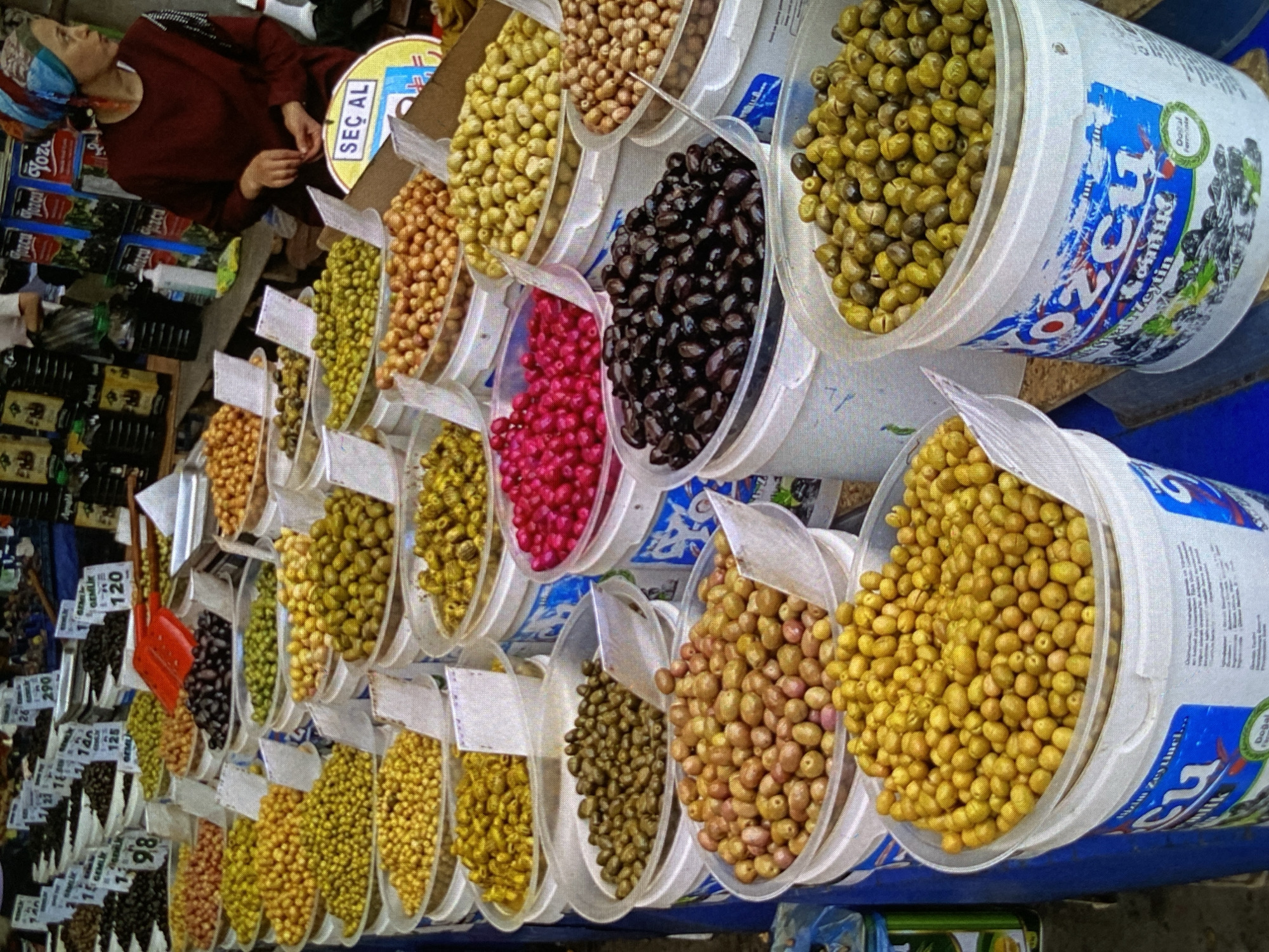 This image depicts a vibrant market stall, likely outdoors, specializing in olives. The main subjects are a vendor and a diverse display of various olive types.
In the upper left, a person, possibly the vendor, is seen from the chest up. They wear a deep maroon long-sleeved top and a colorful headscarf patterned in blue, orange, and white. Their gaze is directed downwards, and their hands are visible, suggesting they are attending to the stall or a customer.
The primary focus of the scene is the extensive array of olives presented in numerous clear plastic containers and larger white buckets. The olives vary significantly in color, including bright green, dark black, reddish-brown, and a unique magenta hue. These are arranged in neat rows across the stall. Many containers have white price tags with numbers such as "98," "140," "26," "125," "290," and "120." A red plastic scoop is placed among the olive containers, indicating how the olives are served.
One large white bucket prominently displays text. A yellow circular sign reads "SEÇ AL," which is Turkish for "Pick and Buy" or "Choose and Take." Other visible Turkish words include "Zeytin" (Olive) and "Doğal" (Natural), along with brand names like "Tozou Yoz," "GEMLIK," and "zcu." These linguistic cues strongly suggest a Turkish origin or influence for the products, even though the provided location context is Bankya, Bulgaria. Below the main olive display, additional products, including bottles that appear to be olive oil, are visible. The scene is brightly lit, indicating daytime.