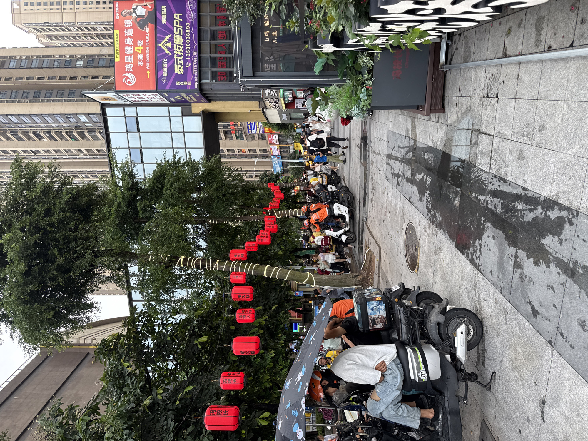 The image depicts a street scene in the Haizhu District of China, characterized by high-rise buildings, pedestrian walkways, and parked electric scooters. Red lanterns are suspended from trees along the left side of the street, adding a festive atmosphere. In the foreground, several electric scooters are parked, some with riders or passengers. One scooter has an open umbrella offering shade to two individuals. A person in light blue jeans and a grey long-sleeved shirt is seated on one scooter, facing away from the camera. The pavement appears wet in places, suggesting recent rain or cleaning. To the right, a walkway is populated by pedestrians, some walking in groups, others individually. A large billboard advertising a fitness center and a spa is visible on the side of a building. The environment suggests a busy urban commercial area.