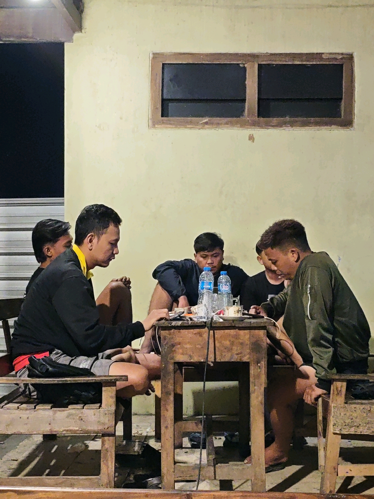 The image shows four young men gathered around a rustic wooden table outdoors, likely in the city of Ngasem, Indonesia. The setting appears to be a casual gathering, possibly in a backyard or a communal area. The men are seated on wooden benches and chairs.
One man, wearing a yellow-collared black jacket and grey shorts, is looking towards the table and appears to be interacting with something on it. Another man, in a black long-sleeved shirt, is also looking at the table. To the right, a man in a green jacket is seated, also focused on the table. Another man, mostly obscured, is seated to the left.
On the table, several items are visible, including two bottles of water, what appear to be small containers or wrappers, and possibly electronic devices or cords. The table itself is worn and weathered, suggesting it is a frequently used piece of furniture.
The background consists of a light-colored wall with a dark, rectangular window featuring wooden framing. The lighting suggests it might be late evening or night, with artificial light illuminating the scene from above. The overall atmosphere is one of relaxed camaraderie.