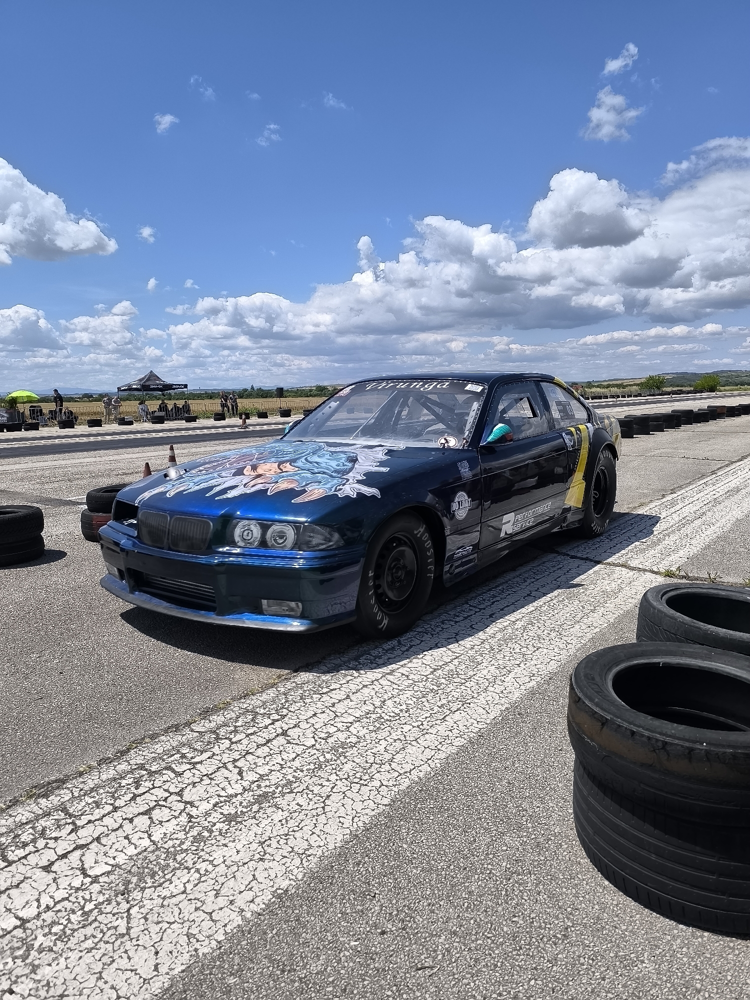 A modified dark blue BMW E36 coupe, customized with a large blue dragon graphic on the hood and various sponsor decals on its sides and windshield, is parked on a paved track under a partially cloudy blue sky. The car has racing tires and a roll cage visible through the windows. In the background, several tents, race barriers, and distant trees are visible, suggesting a racing or event venue. The ground shows cracked white paint lines and tire marks. Stacked tires are visible to the right of the car. The scene appears to be an outdoor event at a race track.