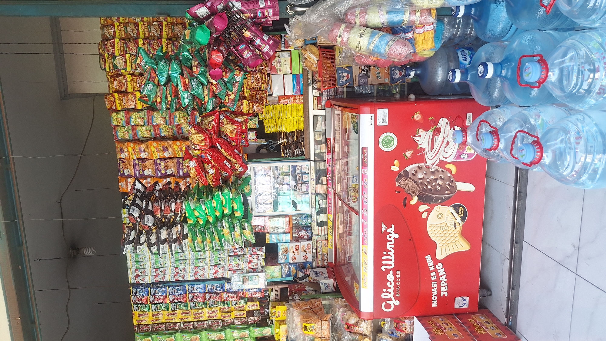 The image captures the interior of a small shop in Wadas, Indonesia, filled with various goods. Racks and walls are densely stocked with an assortment of snacks, candies, and instant noodles, many in colorful packaging. A red ice cream freezer with "Glico Wings" branding is positioned prominently, with bottles of "Aqua" brand water stacked on top. The shop also sells yarn. The scene suggests a typical local convenience store catering to everyday needs in the community of Wadas.