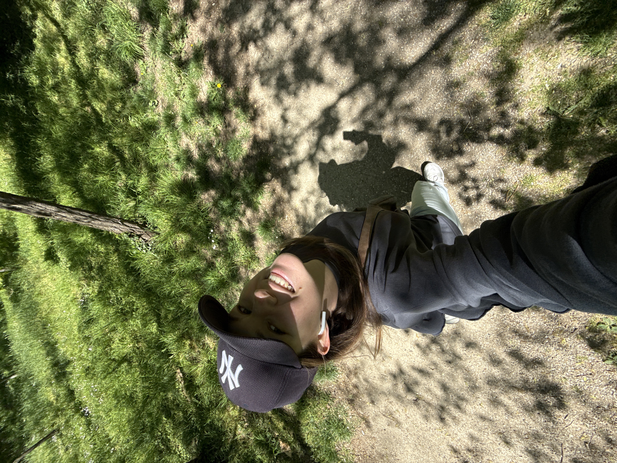 A woman wearing a black New York Yankees baseball cap, a dark grey sweatshirt, and white sneakers is standing on a gravel path partially shaded by trees. Grass is visible to the left. The photo appears to be taken from a low angle, looking up at the subject. The shadow of the person taking the photo can be seen on the gravel path. The location appears to be a park or green space. I am unable to verify the location in the city of Vozdovac Urban Municipality, Serbia from the information provided.