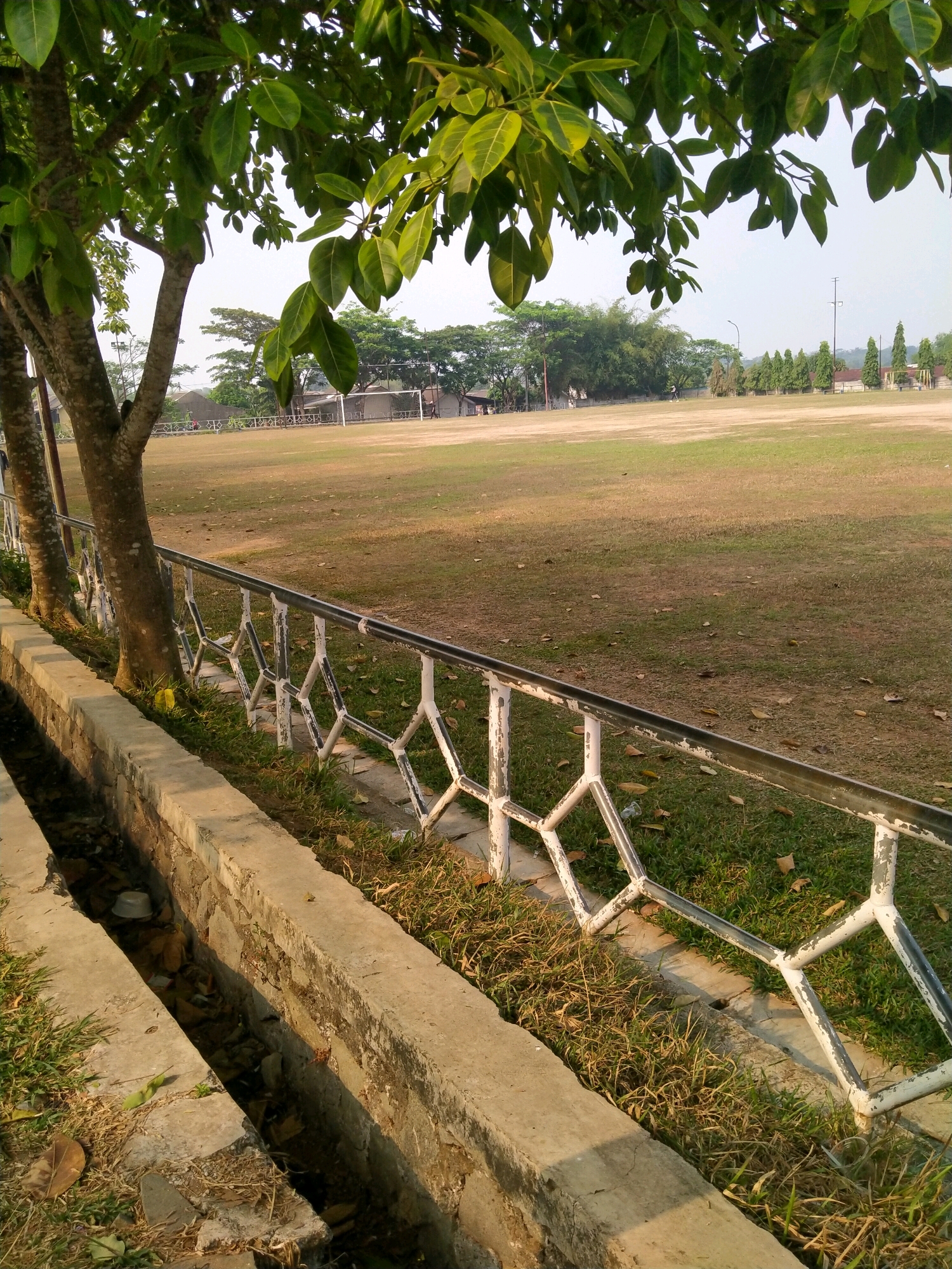 A medium shot captures an outdoor scene in Munjul, Indonesia, likely a sports field or park, bathed in sunlight. In the foreground, a concrete drainage ditch runs along the bottom of the image, filled with debris and some water. Adjacent to the ditch is a low stone wall topped with a white metal fence featuring a diamond pattern. Two trees, their bark textured and greyish, stand near the left edge, partially obscuring the view. Overhanging branches and green leaves from a large tree dominate the upper left portion of the frame.
The background opens up to a vast, brownish-green field, appearing dry and sparsely covered with grass, dotted with fallen leaves. Beyond the field, a line of trees forms a backdrop, with some smaller, manicured trees in a row on the right side of the field. In the distance, a soccer goal with a white net is visible on the left side of the field, and several buildings with reddish roofs are present. The sky above is a pale, hazy blue. The overall impression is one of a warm, sunny day in a somewhat neglected but functional outdoor public space.
