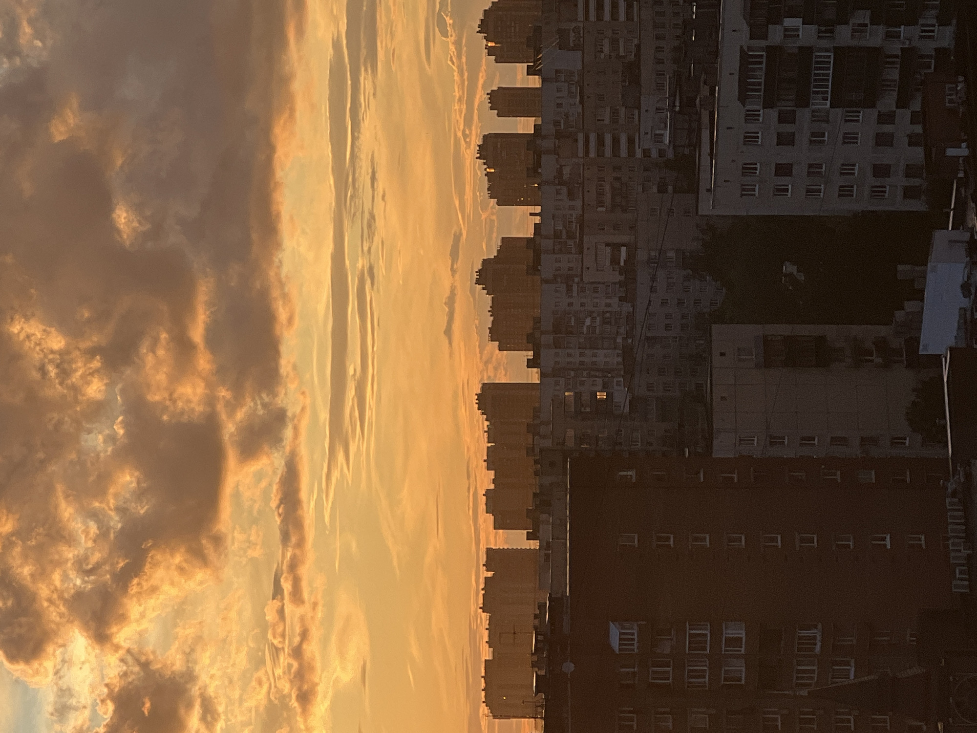 The image shows a cityscape at sunset. The left side of the frame is dominated by a dramatic sky filled with clouds illuminated by the setting sun, casting warm orange and yellow hues. Streaks of lighter clouds crisscross the sky, suggesting movement and atmospheric depth.
On the right side of the frame, a dense collection of apartment buildings is silhouetted against the sky. The buildings are of varying heights and architectural styles, with many windows visible. Some windows are illuminated, indicating that the residents are home. The buildings are packed closely together, creating a sense of urban density.
The composition is split vertically, with the sky occupying the left half and the city the right half. This juxtaposition highlights the contrast between the natural beauty of the sunset and the man-made environment of the city.
The time of day is clearly sunset, indicated by the warm, low-angled sunlight and the dramatic cloud formations. The weather appears to be clear, with some cloud cover. The location is urban, with many tall buildings. The mood conveyed is one of tranquility and the beauty of a natural phenomenon occurring within a bustling city. There is no visible text in the image.