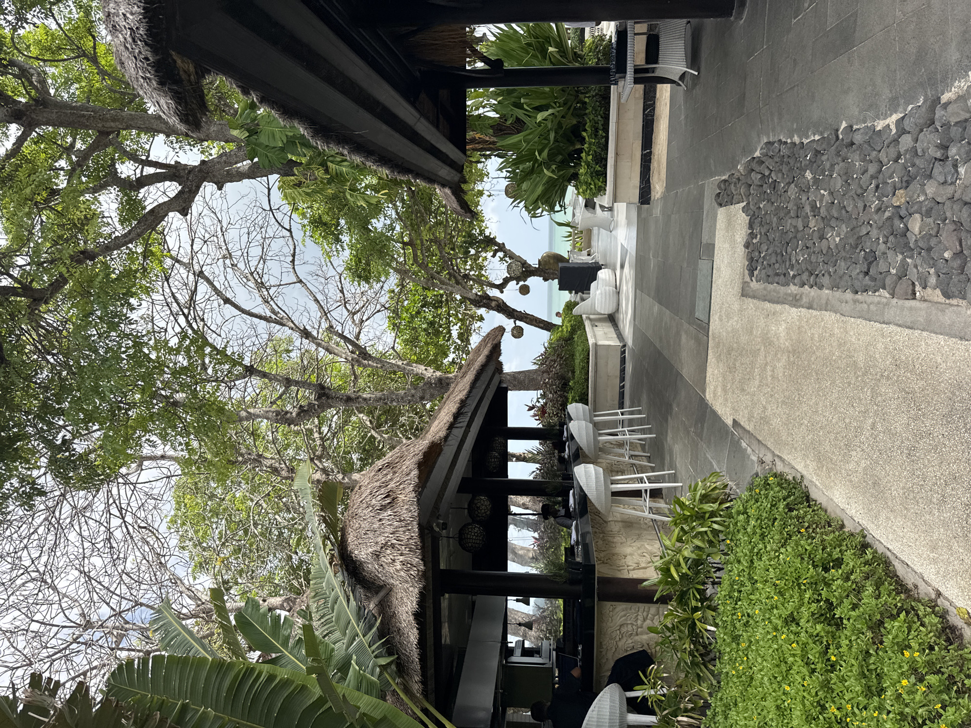 The image shows an outdoor restaurant or bar setting in Kuta Selatan, Indonesia. The location features a thatched-roof structure, likely a bar, with pendant lighting hanging from the eaves. White bar stools line the counter. Lush tropical foliage surrounds the structure, including banana plants and other greenery. A paved walkway leads away from the bar, bordered by decorative rock features. In the distance, there is a glimpse of the ocean. The scene appears tranquil and tourist-oriented.