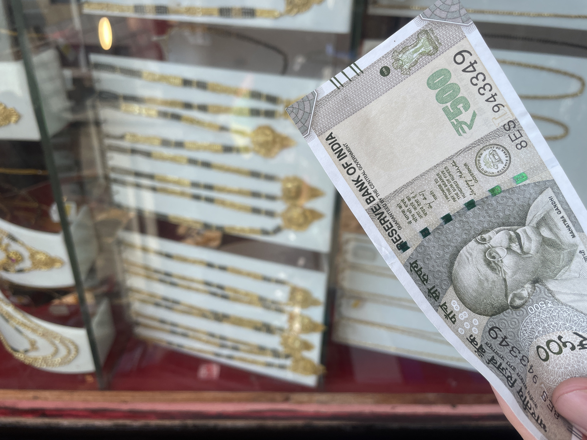 A human hand holds a crisp 500 Indian Rupee banknote in the foreground, angled upwards and to the right. The banknote displays the portrait of Mahatma Gandhi and the serial number "8ES 943349". The text "RESERVE BANK OF INDIA" and "पांच सौ रुपये" are clearly visible. In the blurred background, behind the banknote, is a jewelry shop display window in Kolhapur, India. Multiple gold necklaces, appearing to be Mangalsutras with black beads and ornate gold pendants, are showcased on white stands across at least two glass shelves. Additional multi-strand gold necklaces are visible on the upper shelf. Red framing elements are discernible on the left side of the display. The scene depicts a financial transaction or consideration occurring in front of a jewelry retail environment.