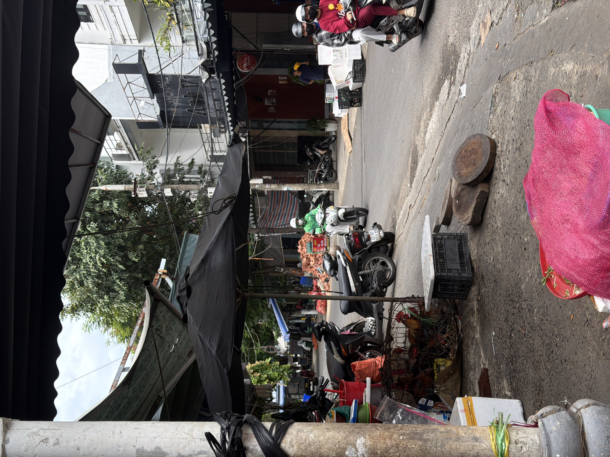 The image depicts a street scene in a market area of Đà Nẵng, Vietnam, likely a commercial district. In the foreground, a weathered concrete pillar wrapped with black plastic tape stands on the left. Adjacent to it, various market goods are displayed, including a wire cage containing live chickens, red buckets, a white cooler box, and a pile of pink netting. Two motorbikes are parked in the middle ground. On the right side of the road, a person is riding a motorbike with a passenger behind them, both wearing face masks. The road is paved, and there are buildings lining the street, some with awnings and balconies. Market stalls and awnings are visible throughout the scene, suggesting active commerce. The overall environment appears to be an outdoor market on a cloudy day.